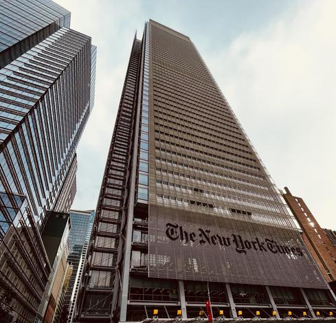 A color photograph in square aspect ratio of the New York Times tower, a tall skyscraper in Manhattan. The photo captures the entire height of the tower as it tapers slightly against a bright, light blue sky with scattered white clouds. The building's facade is notable for its steel-and-glass curtain wall, and a large, stylized "The New York Times" logo is prominently displayed in dark lettering across the lower portion of the visible exterior, set against the light vertical louvers that partially screen the glass. The composition is framed by the adjacent buildings, one of which is a much starker, dark glass structure on the left that reflects the sky and the surrounding city. The bottom of the image shows a high street level, with the roof of the building’s entry visible. The street and sidewalks are not visible.