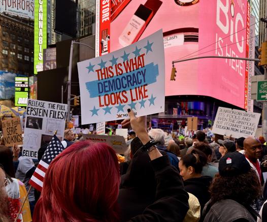 A color photograph of the recent No Kings protest in Times Square, Manhattan. The square is packed full of thousands of people. Many are holding up signs and American flags. Notable signs include: We the people serve no kings. Release the full Epstein files! A nation can survive its fools, and even the ambitious. But it cannot survive treason from within. - Cicero. The most prominent sign is the center and focal point of the image, it reads “This is what democracy looks like. No kings.” The sign is held by a woman with her right hand. Her nails have dark nail polish, her hair is dyed a deep red, and she is wearing a black wind breaker jacket. The digital billboard ads light up the scene from above the protestors.