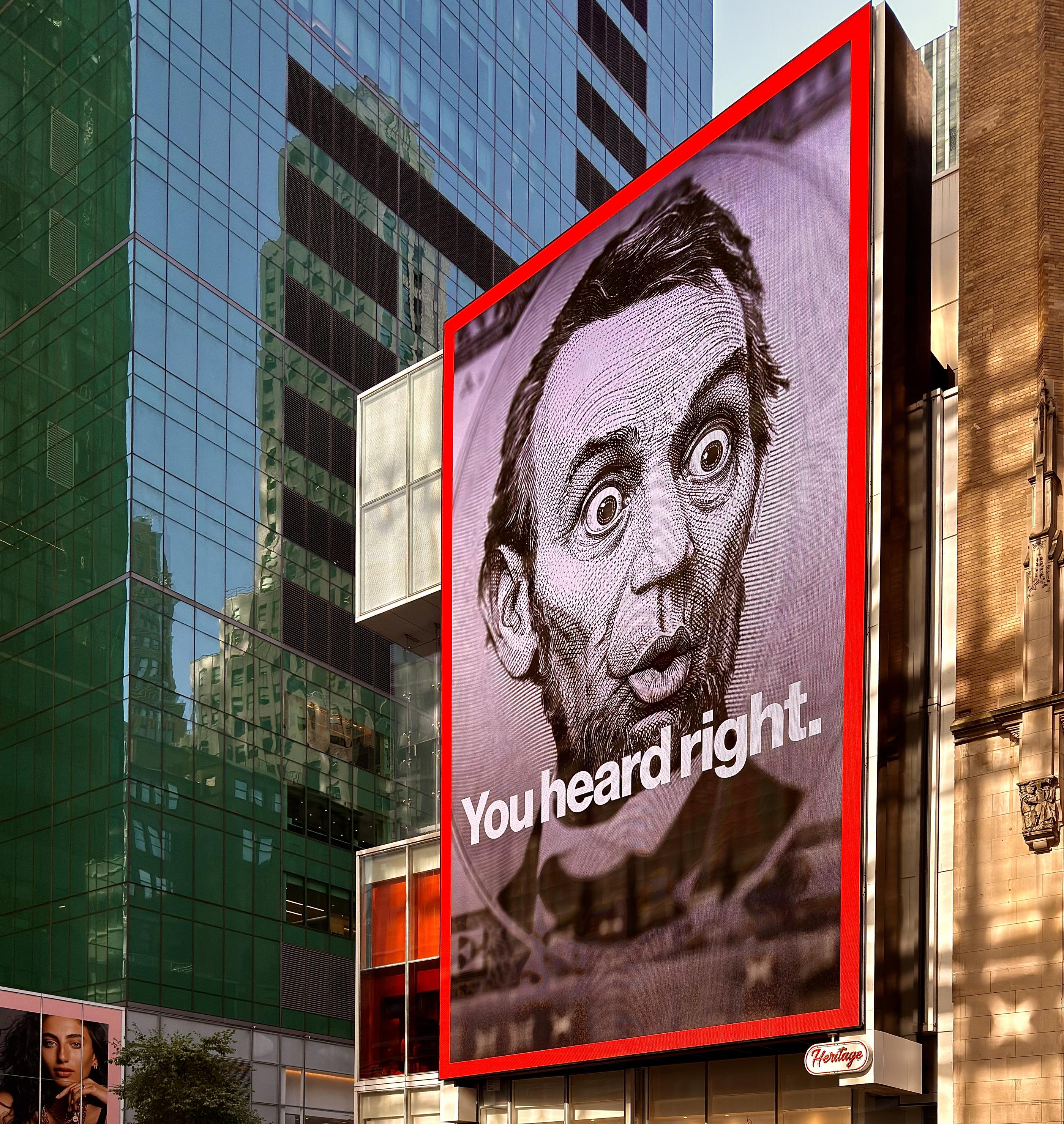 A color photograph in square aspect ratio of a very large, vertically oriented, digital billboard along 42nd Street near the intersection with Broadway in midtown Manhattan. The screen is displaying a close up depiction of Lincoln on the $5 bill with an edited image showing his eyes open wide in shock and his lips pursed in disbelief. Large white text is beneath his face that reads “You heard right.” There is a red border around the screen. Late afternoon sunlight is filtering onto a tan brick building behind the signature, and a deep blue colored glass building fills the rest of the frame.