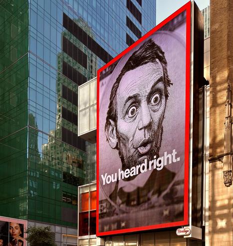 A color photograph in square aspect ratio of a very large, vertically oriented, digital billboard along 42nd Street near the intersection with Broadway in midtown Manhattan. The screen is displaying a close up depiction of Lincoln on the $5 bill with an edited image showing his eyes open wide in shock and his lips pursed in disbelief. Large white text is beneath his face that reads “You heard right.” There is a red border around the screen. Late afternoon sunlight is filtering onto a tan brick building behind the signature, and a deep blue colored glass building fills the rest of the frame.