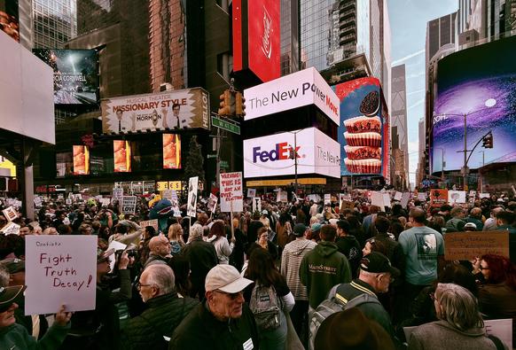 A color photograph of the No Kings protest as it began to gather momentum in Father Duffy Square at 10:30am in Manhattan. A dense crowd of thousands of people fills the square and the surrounding streets. The crowd is holding a multitude of diverse signs and flags that voice their opposition to the Trump administration. Signs with phrases like "ICE out of NYC," "fight fascism," "we love America," and "No Kings Since 1776" are visible above the heads of the crowd, contrasting with the commercial advertisements of the surrounding billboards. In the lower left corner a man wearing a dark colored baseball cap holds a white sign with red lettering that reads “Dentists Fight Truth Decay!”. The digital billboard are advertising Broadway shows, FedEx, Oreo and Reese’s cookies. The sky is seen between the buildings as a light blue color. 