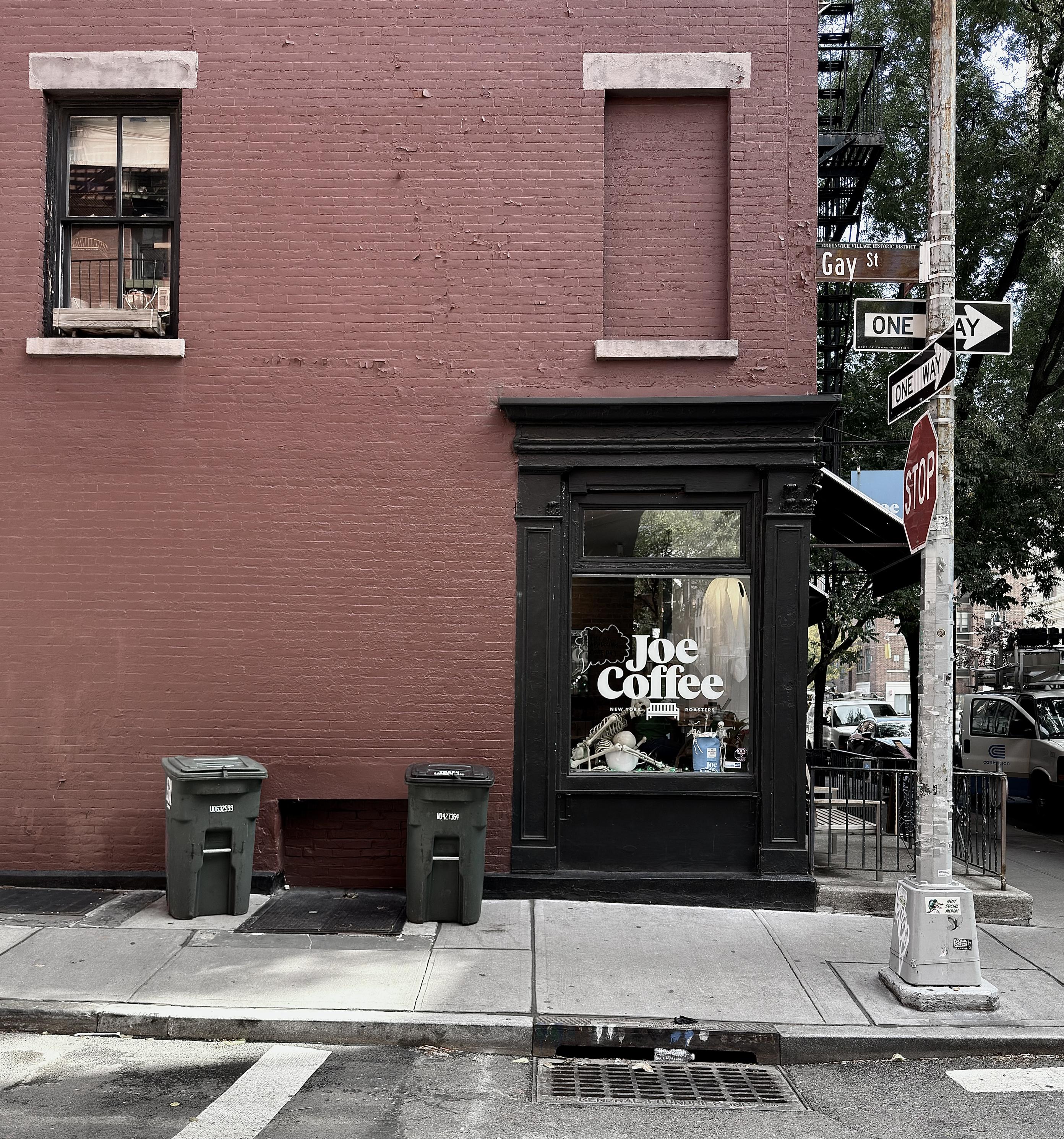 A color, but desaturated, photograph of the street scene along Waverly Place at the intersection with Gay Street in the West Village neighborhood of Manhattan. In the center of the frame is the Joe Coffee shop located on the corner. The building wall is a striking, deep reddish-brown brick, contrasting with the dark black and brown trim of the storefront and the windows. The coffee shop entrance is recessed and framed in a decorative molding with a large window displaying the white "Joe Coffee" signage. Above the entrance, there's a tall, rectangular recess in the brickwork where a window has been filled in, and to the left, a single-paned vertical window with a slightly worn black frame. The lower part of the brick wall is visible, with two standard dark green urban trash/recycling bins sitting on the sidewalk in front of it. The street scene to the right includes a metal pole holding a street sign assembly. The signs clearly point to "Gay St" and indicate that both streets are ONE WAY. A red STOP sign is also mounted on the pole. The sidewalk and street are concrete, with a metal drain grate visible near the curb. In the background, beyond the signs and pole, there is a glimpse of other parked vehicles, street activity, and the muted green of trees. Taken early afternoon on a partly cloudy day.
