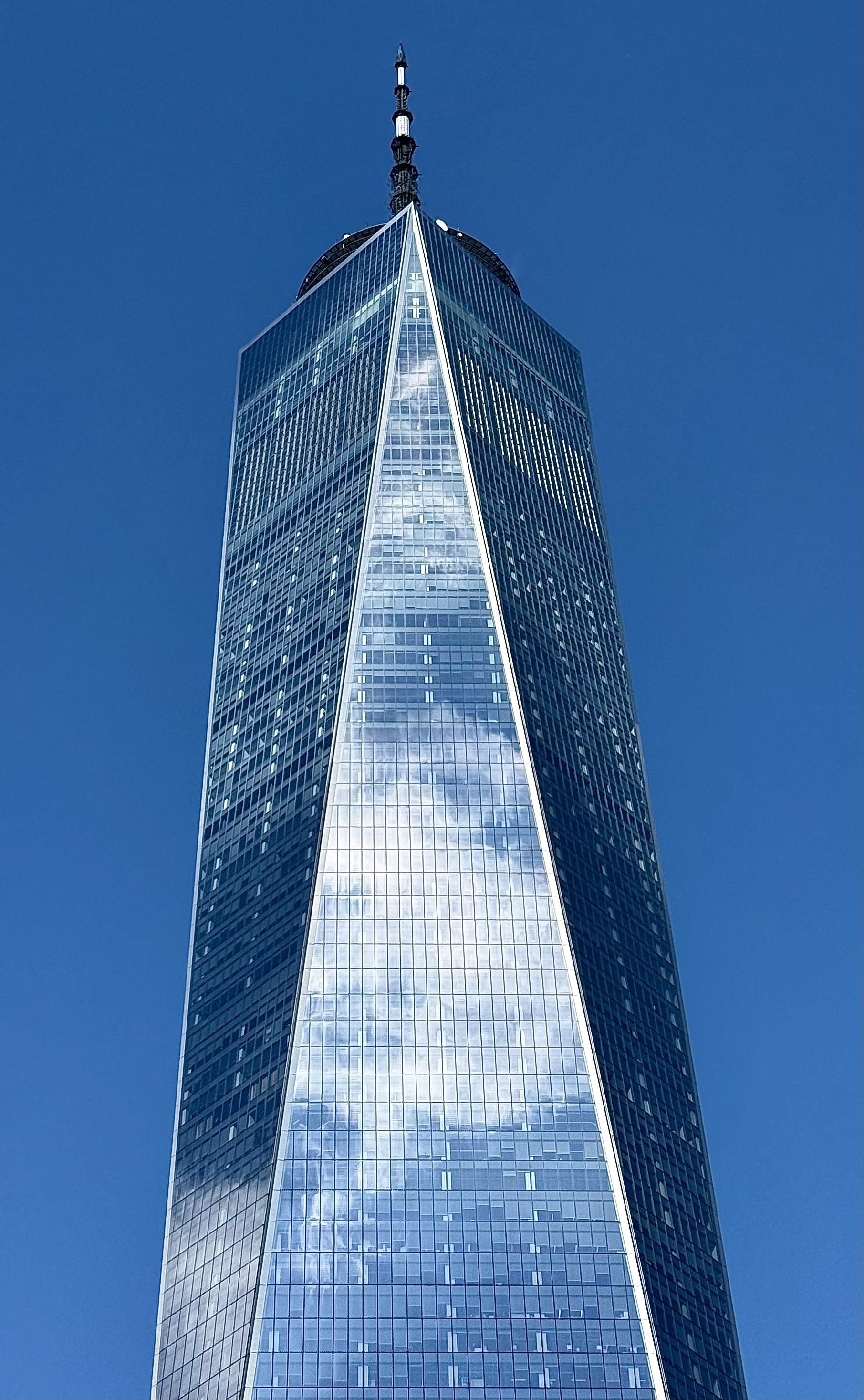 A color photograph in portrait orientation of One World Trade Center in New York City. The striking, angular skyscraper dominates the frame against a clear, vibrant blue sky. The building's glass facade reflects the sky and a scattering of white clouds, creating a brilliant, shimmering effect, especially along the central, triangular section that tapers toward the top. The image emphasizes the sheer height and sharp architectural lines of the tower. The photo is tightly cropped, focusing on the upper two-thirds of the building and culminating at the spire at its apex. The light is bright, highlighting the reflective surfaces and modern design of the structure.