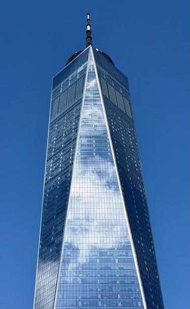 A color photograph in portrait orientation of One World Trade Center in New York City. The striking, angular skyscraper dominates the frame against a clear, vibrant blue sky. The building's glass facade reflects the sky and a scattering of white clouds, creating a brilliant, shimmering effect, especially along the central, triangular section that tapers toward the top. The image emphasizes the sheer height and sharp architectural lines of the tower. The photo is tightly cropped, focusing on the upper two-thirds of the building and culminating at the spire at its apex. The light is bright, highlighting the reflective surfaces and modern design of the structure.
