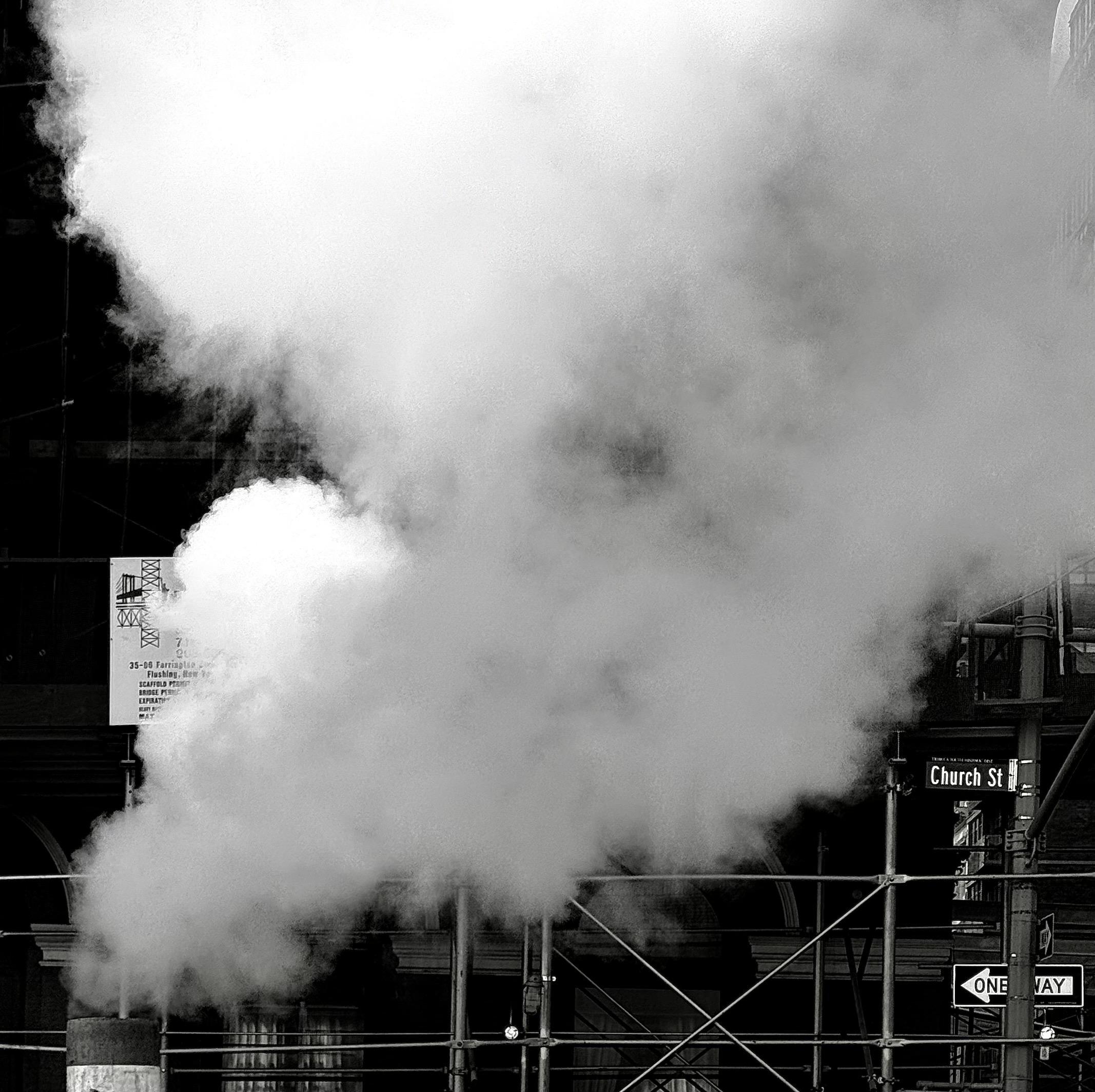A black and white street photograph capturing a large plume of steam rising vertically and billowing outwards. The steam dominates the center and upper portion of the frame, creating a soft, ethereal texture against the dark, gritty background. The origin of the steam is low in the frame, a utility vent, and is partially obscured by the framing of the image. The background consists of dark, silhouetted building facades and silver construction scaffolding. Several street signs are visible on the right, including a sign reading "Church St" and a sign below it with a white arrow indicating "ONE WAY." A partial, lighter-colored sign is partially visible with obscured writing through the steam on the left. The scene is high contrast, and the overall mood is dramatic and characteristic of an urban environment, emphasizing texture and the stark contrast between light and shadow.