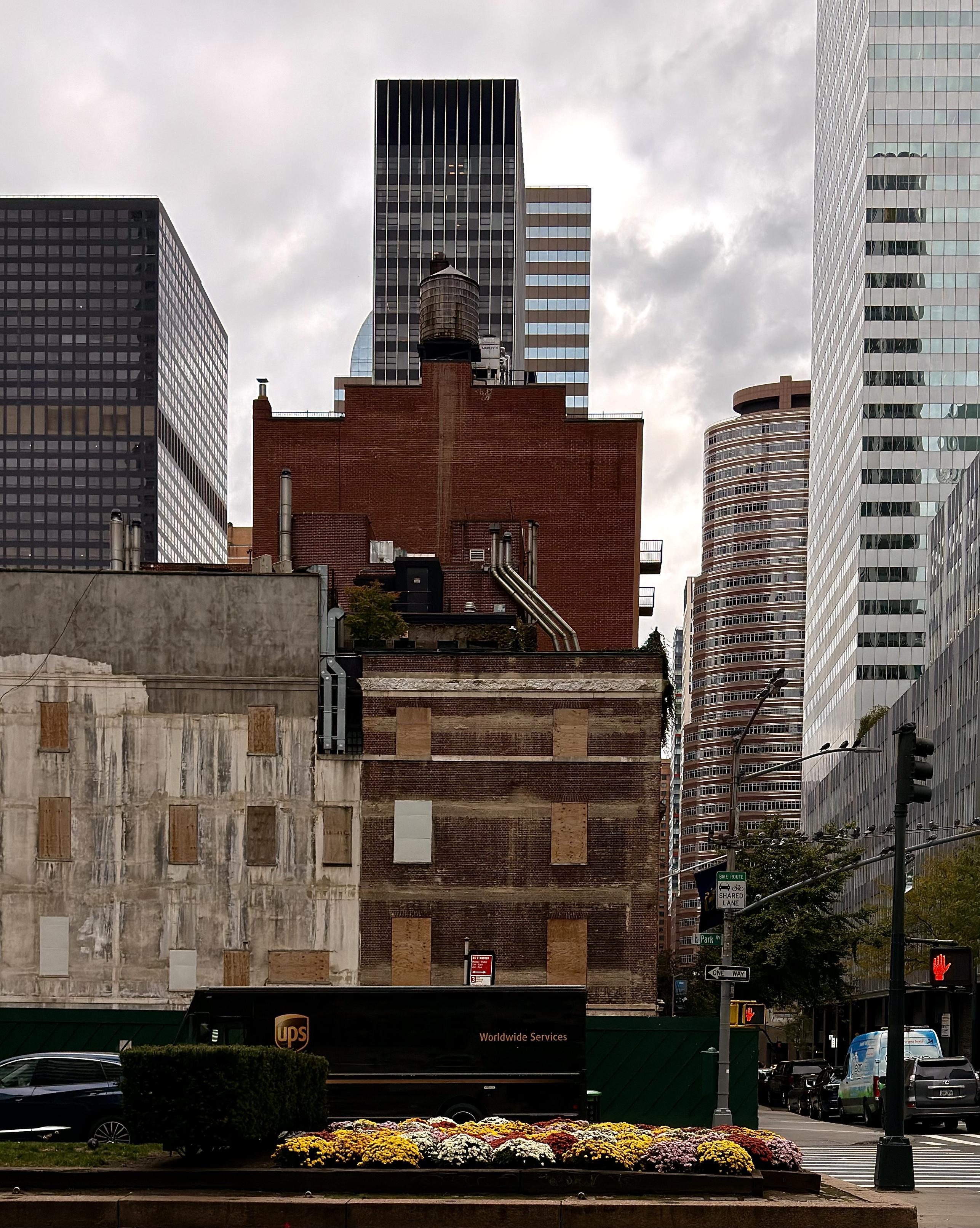 A color photograph in portrait orientation of the street scene along Park Avenue in midtown Manhattan on a cloud morning. In the foreground, a low brick building stands out with a classic water tower perched on its roof, flanked by taller, modern glass and steel skyscrapers that dominate the background. The immediate street level features a raised, well-kept flower bed filled with yellow, orange, and red chrysanthemums, providing a burst of color against the dark tones of the surrounding scene and a brown UPS delivery truck waiting in traffic. The overall composition emphasizes the contrast between old and new architecture, with the central brick structure showing signs of aging, including boarded-up windows on both its beige and darker brick sections. The tall buildings behind it include one dark and geometric on the left, and a cylindrical, modern skyscraper on the right. A traffic light and street signs are visible on the right, with a red ‘Don’t walk’ hand signaling pedestrian traffic. Beyond the traffic sign there are cars parked along a side street. There are no people, and no movement in the scene.