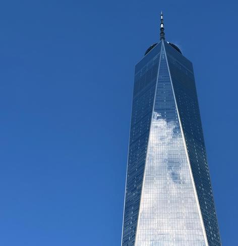 A color photograph in square aspect ratio of One World Trade Center in New York City. The striking, angular skyscraper dominates the frame against a clear, vibrant blue sky. The building's glass facade reflects the sky and a scattering of white clouds, creating a brilliant, shimmering effect, especially along the central, triangular section that tapers toward the top. The image emphasizes the sheer height and sharp architectural lines of the tower. The photo is tightly cropped, focusing on the upper two-thirds of the building and culminating at the spire at its apex. The light is bright, highlighting the reflective surfaces and modern design of the structure.