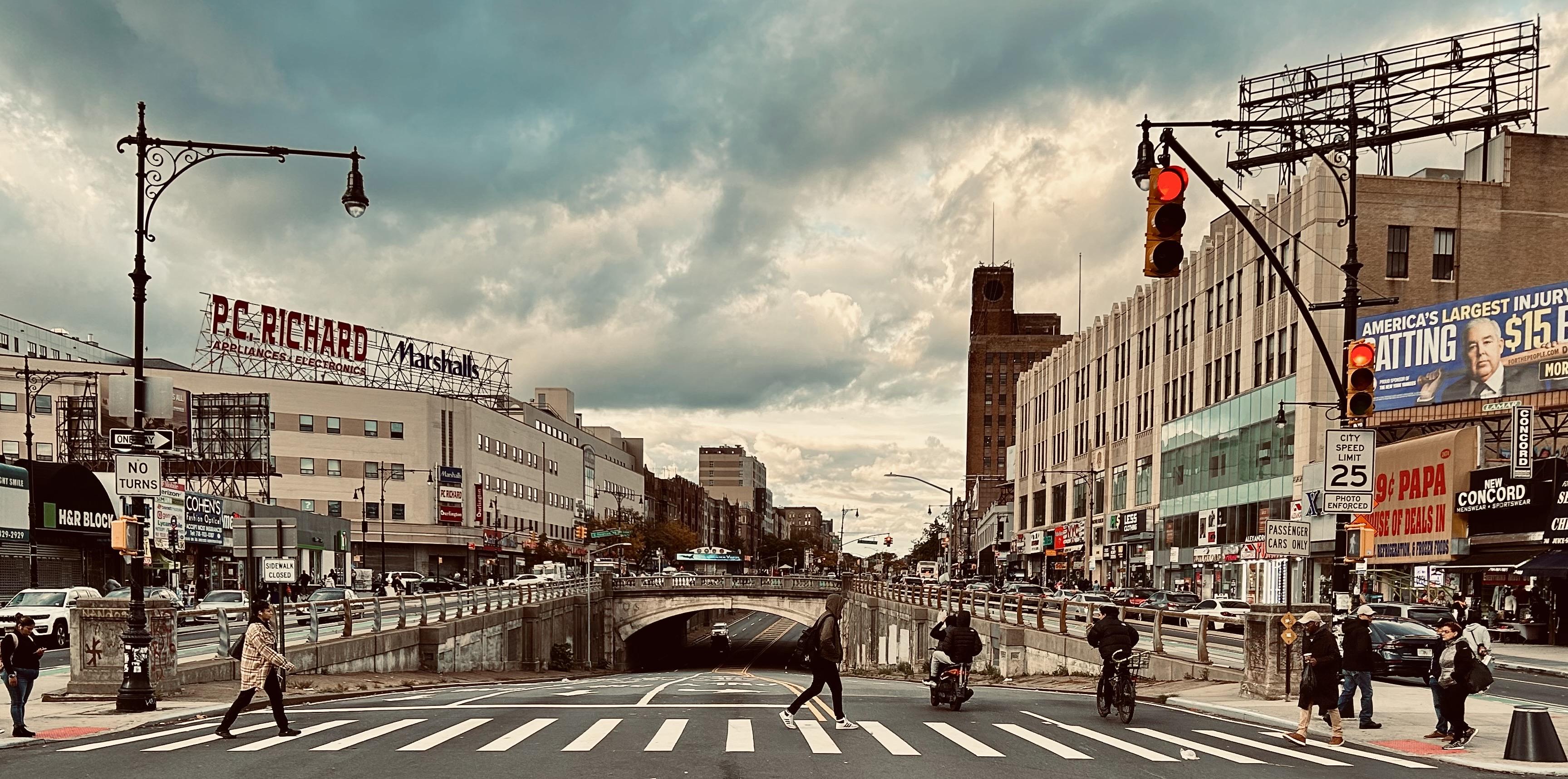 A color photograph in landscape orientation of the street scene along the Grand Concourse at the intersection with Fordham Road. The composition centers on the concourse underpass structure that cuts horizontally across the middle ground. Pedestrians are actively crossing the street in the foreground, using a marked crosswalk; several figures are walking towards the center, one is on a scooter, and another on a bicycle. The lighting is dramatic, with an overcast sky featuring rich, textured clouds that dominate the upper half of the image. The surrounding area is commercial, both sides of the street are lined with multi-story brick and concrete buildings featuring prominent signage. On the left, large billboards read "P.C. RICHARD & SON APPLIANCES, ELECTRONICS" and "Marshalls." On the right, a large illuminated billboard features an advertisement for a personal injury attorney. A traffic light on the right is red. The street is busy with traffic flowing beneath the underpass and along the main street.