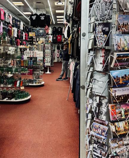 A color photograph in portrait orientation of a souvenir shop in Times Square, Manhattan. The shop has a long, narrow layout with a medium red color carpeted floor that draws the eye toward the back. The walls are densely packed with merchandise. On the left, tiered display shelves hold a variety of small souvenirs, including miniature replicas of landmarks like the Statue of Liberty, and other novelty items. The walls and overhead racks are completely covered in clothing, primarily T-shirts and sweatshirts with "New York City" themes, predominantly in red, white, and black colors. In the center-right of the frame, the owner of the store, a man dressed in a dark jacket and light pants, stands with his open hands against his cheeks while looking in front of himself. A prominent feature in the foreground on the right is a tall, metal rotating postcard rack, filled with dozens of vibrant postcards showcasing various iconic New York City scenes and skylines.