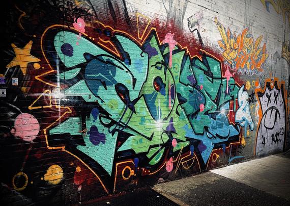 A color photograph in landscape orientation of a vibrant and dynamic graffiti art mural covering a weathered brick wall on a city street. The primary piece of graffiti is a complex "wildstyle" mural dominated by shades of teal, lime green, and dark blue, with bold black outlines and highlights of bright yellow-orange and hot pink dots and accents. The lettering is highly stylized and interlocking, and its energetic, angular shapes give the composition a strong sense of movement. The mural extends across the center and left of the frame, with smaller, older, and less defined tags and pieces visible on the right. In the far right background, a faded white and gray character-style piece is partially visible, offering a stark contrast to the main vibrant work. The photo is taken at street level, looking slightly up and along the wall, which recedes into the distance on the right.