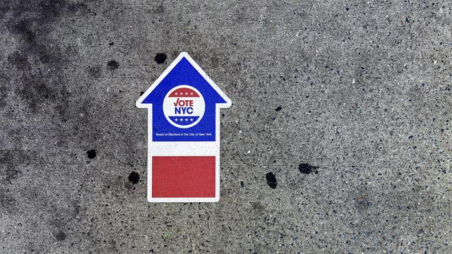 A color photograph in landscape orientation of a sidewalk, taken on an overcast morning in the Upper West Side neighborhood of Manhattan as I was walking to the polls to cast my vote in opposition of the Democratic mayoral frontrunner. The focal point of the image is an eye-level, close-up shot of a "VOTE NYC" sticker placed on the rough, dark gray concrete surface. The sticker is shaped like a house with an upward-pointing arrow for a roof. The upper, arrow-shaped portion is blue and contains a white circle with red trim on top, blue trim on bottom, that reads "VOTE NYC". The word VOTE is red text with the letter V stylized to look like a check mark ✔️. The word NYC is blue text. The trim on top and bottom of the circle each have four white stars in a line. Below the main logo, in smaller text, it reads "Board of Elections in the City of New York." The lower, rectangular portion of the sticker is solid red. The surrounding concrete pavement is speckled with various dark stains. Two black speckles on each side of the lower portion of the sticker, and two more on the left side of the upper portion of the sticker. The concrete is stained a dark grey color on the upper left corner of the frame. The image was captured in the shade of a building so there are no light casts or shadows in the scene.
