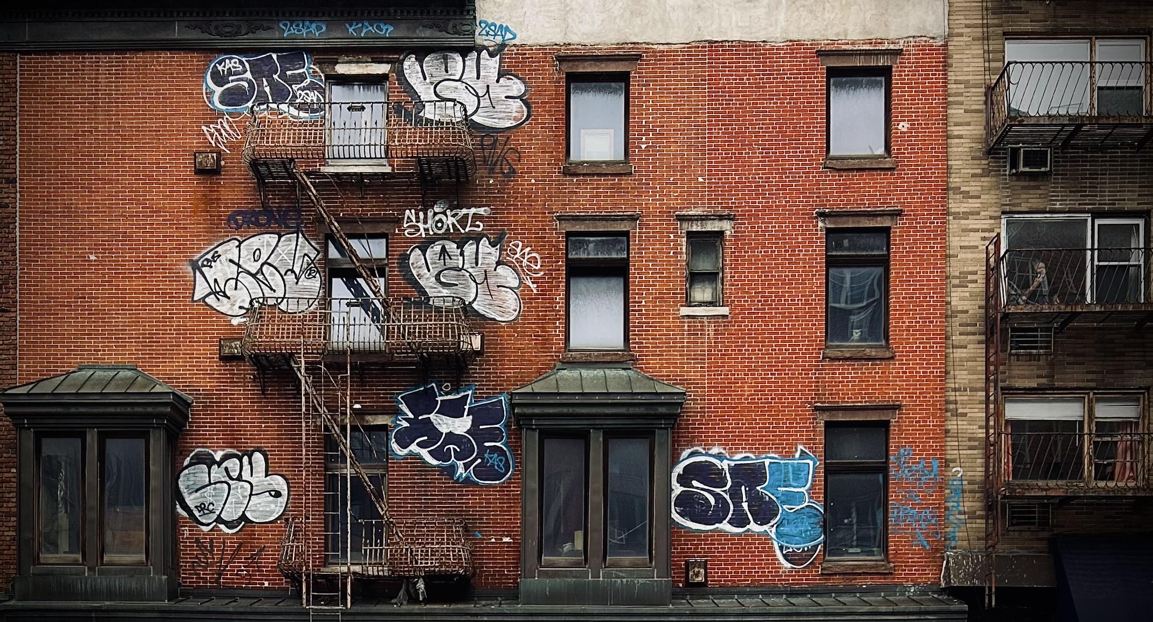 A color photograph in landscape orientation of the screen scene in the Chelsea neighborhood of Manhattan captures the gritty facade of two brick apartment buildings. It is an overcast morning so the mood is dim, and there is no light casting or shadows. The main building on the left consumes 75% of the frame, and is composed of a reddish-brown brick, with several windows, some large, double-paned ones on the first floor, and smaller, single-pane windows on the upper floors, featuring dark window frames and ledges. In the center floor a very small window sits to the right of another window, likely a bathroom. A rusty, metal fire escape zigzags down the left side of the building, connecting several small metal balconies outside upper-story windows. The most prominent feature is the extensive, stylized graffiti sprayed across the brickwork behind and next to the fire escape structure. Large, bubble-letter tags in white, black, and blue cover much of the left and center portions of the wall. Several large, illegible pieces are visible near the upper windows and below the main fire escape landing. To the right, a neighboring building with lighter, tan and brown brick and white framed windows behind metal balconies. 