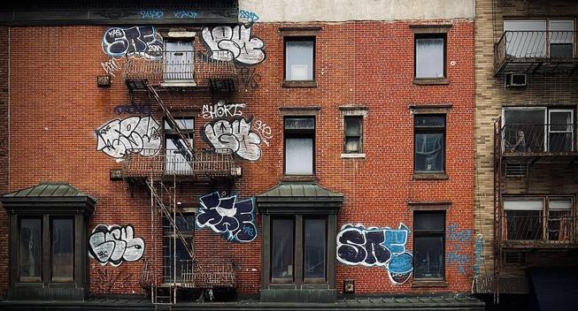 A color photograph in landscape orientation of the screen scene in the Chelsea neighborhood of Manhattan captures the gritty facade of two brick apartment buildings. It is an overcast morning so the mood is dim, and there is no light casting or shadows. The main building on the left consumes 75% of the frame, and is composed of a reddish-brown brick, with several windows, some large, double-paned ones on the first floor, and smaller, single-pane windows on the upper floors, featuring dark window frames and ledges. In the center floor a very small window sits to the right of another window, likely a bathroom. A rusty, metal fire escape zigzags down the left side of the building, connecting several small metal balconies outside upper-story windows. The most prominent feature is the extensive, stylized graffiti sprayed across the brickwork behind and next to the fire escape structure. Large, bubble-letter tags in white, black, and blue cover much of the left and center portions of the wall. Several large, illegible pieces are visible near the upper windows and below the main fire escape landing. To the right, a neighboring building with lighter, tan and brown brick and white framed windows behind metal balconies. 