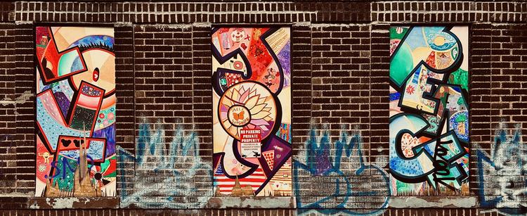 A color photograph in landscape orientation of a street art installation in the Bay Ridge neighborhood of Brooklyn. The scene consists of a section of a dark brick wall featuring three rectangular, vertical murals installed within former window openings. Each mural is filled with vibrant, overlapping abstract and illustrative elements in a dynamic, collage-like style. The brickwork is dark brown/red and runs across the top and between the murals, while the bottom section of the wall below the murals shows signs of blue and light-colored graffiti tags that are partially washed out or faded. From left to right the murals spell out the words: love, joy, peace. The leftmost mural primarily uses tones of red, blue, purple, and yellow/tan, featuring bold, curved, and angular shapes. The center mural is dominated by reds, oranges, and pinks, and prominently features a large, stylized sunflower with a butterfly and the text ‘Sally Sunshine’ wrapped around it. A small red and white sign is visible over the flower, stating "NO PARKING PRIVATE PROPERTY. Violators will be towed away at owner’s expense." The rightmost mural is characterized by vivid greens, teals, and blues. All three murals have incorporated within the letter shapes many smaller details such as floral and technological elements. It is day time and sunlight is illuminating the scene, but not light casts or shadows are visible in the scene.
