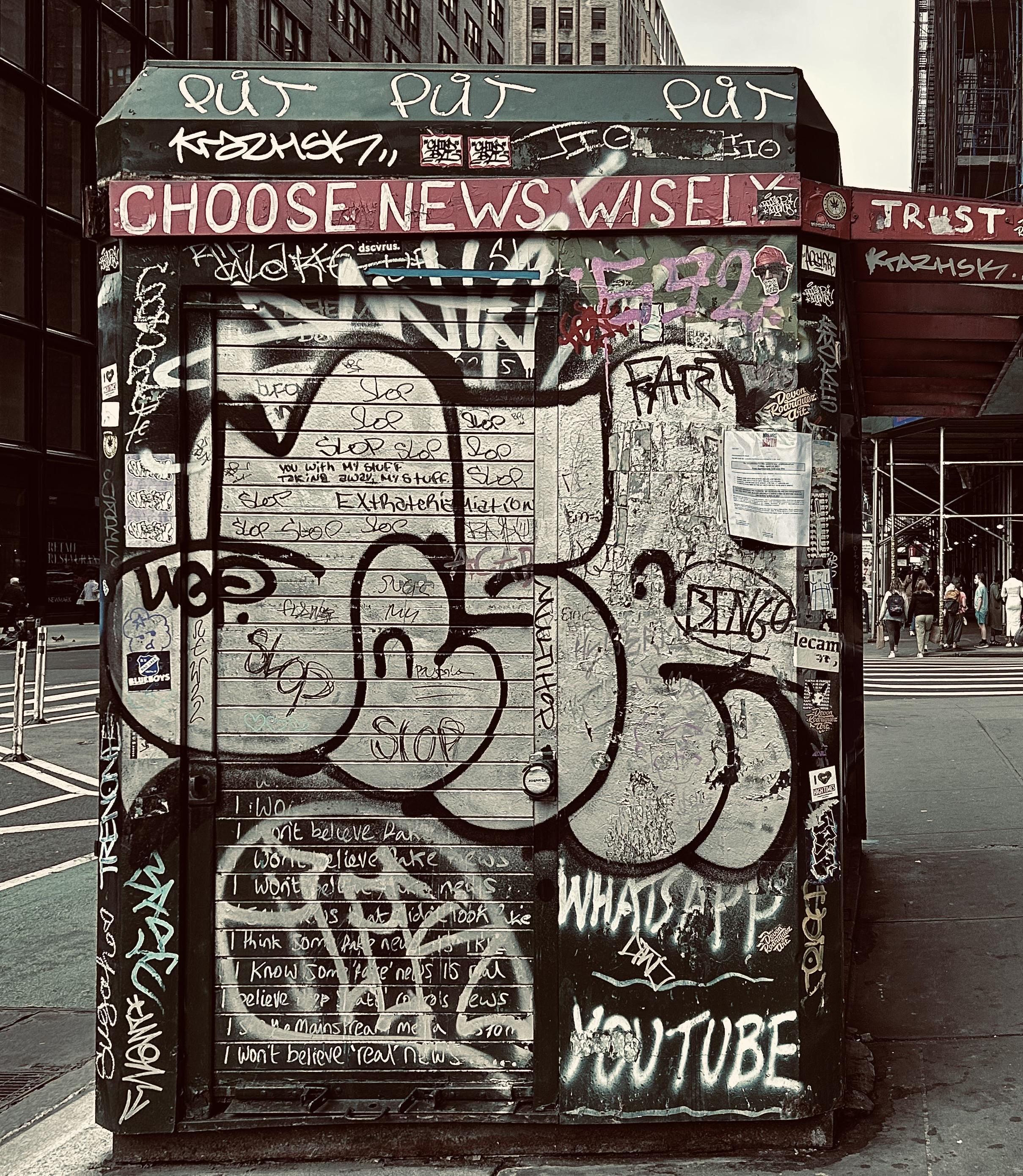 A color, desaturated photograph of the street scene along seventh avenue at the intersection with 28th street in the Chelsea neighborhood of Manhattan. It is day time and party cloudy. The focal point is a shuttered dark green newsstand kiosk. The entire exterior wall of the structure is covered in dozens of graffiti tags. The most notable is white text layered across a red strip of paint near the top of the wall that reads, ‘CHOOSE NEWS WISELY. NO TRUST’. Beyond the kiosk the crosswalk and street are visible to the left, a small group of people are visible on the right, gathered across the street waiting to cross. Brick apartment buildings fill the rest of the frame on each side of the frame. 