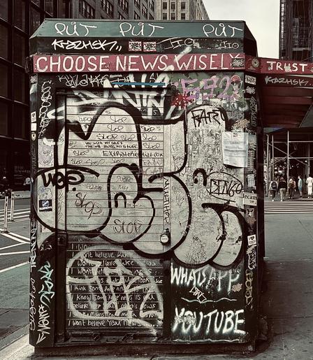 A color, desaturated photograph of the street scene along seventh avenue at the intersection with 28th street in the Chelsea neighborhood of Manhattan. It is day time and party cloudy. The focal point is a shuttered dark green newsstand kiosk. The entire exterior wall of the structure is covered in dozens of graffiti tags. The most notable is white text layered across a red strip of paint near the top of the wall that reads, ‘CHOOSE NEWS WISELY. NO TRUST’. Beyond the kiosk the crosswalk and street are visible to the left, a small group of people are visible on the right, gathered across the street waiting to cross. Brick apartment buildings fill the rest of the frame on each side of the frame. 