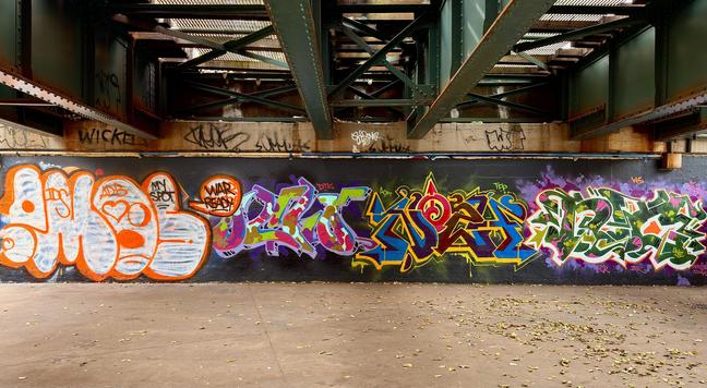 A color, wide-angle photograph in letterbox orientation of a colorful graffiti mural painted atop a black wall beneath a large, elevated steel railway structure for the NYC subway tracks just south of Yankee Stadium in the Bronx. The underpass has large, dark green, crisscrossing steel beams and girders overhead. The floor in the foreground is a light brown, rough concrete scattered with dried brown and yellow leaves. The mural itself is a complex arrangement of multiple large graffiti tags created in different styles and color palettes. From bold orange and white bubble letters to purple, pink, and bright green color schemes featuring sharp, interlocking letters, followed by a piece dominated by deep brown, yellow, and bright blue in an intricate wildstyle. Thw concrete support wall directly above the mural is a stained light brown color and features various smaller, dark tags and scribbles. It is an overcast morning and sunlight is filtering down into the scene from open metal grating on the track structure above.