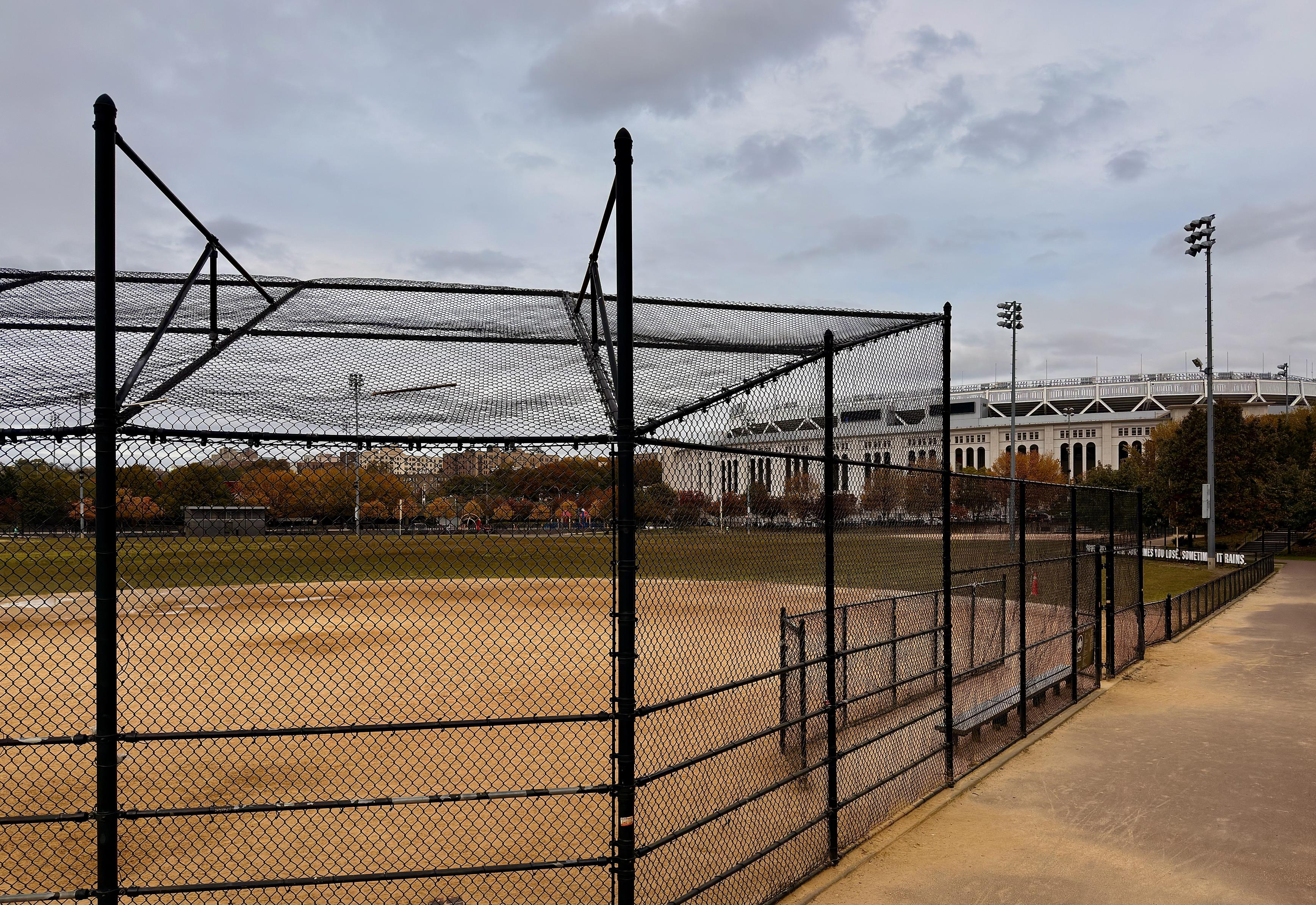 A color photograph of the view from the side of a baseball diamond at Heritage Field, with the prominent structure of Yankee Stadium visible in the distance under a cloudy, overcast morning sky. In the foreground, a tall black chain-link fence, supported by black vertical posts, separates the walking path running along the right side of the frame from the baseball field. The field is primarily a dusty tan color with a faint backstop netting and framework visible closer to the left. A set of black metal spectator benches is visible just inside the fence line to the right. The middle ground features the expansive baseball diamond, which is void of people. Beyond the outfield, a line of trees displays autumn foliage in muted shades of gold and russet. A playground is visible at the far side of the field. Cars parked along the street border the park. Dominating the background is the distinct, white-pillared upper facade of Yankee Stadium. Its grandstands and upper decks are visible above the surrounding structures. The scene is illuminated by tall stadium lights for the smaller field and others for the stadium.