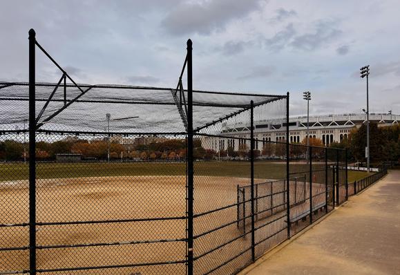 A color photograph of the view from the side of a baseball diamond at Heritage Field, with the prominent structure of Yankee Stadium visible in the distance under a cloudy, overcast morning sky. In the foreground, a tall black chain-link fence, supported by black vertical posts, separates the walking path running along the right side of the frame from the baseball field. The field is primarily a dusty tan color with a faint backstop netting and framework visible closer to the left. A set of black metal spectator benches is visible just inside the fence line to the right. The middle ground features the expansive baseball diamond, which is void of people. Beyond the outfield, a line of trees displays autumn foliage in muted shades of gold and russet. A playground is visible at the far side of the field. Cars parked along the street border the park. Dominating the background is the distinct, white-pillared upper facade of Yankee Stadium. Its grandstands and upper decks are visible above the surrounding structures. The scene is illuminated by tall stadium lights for the smaller field and others for the stadium.