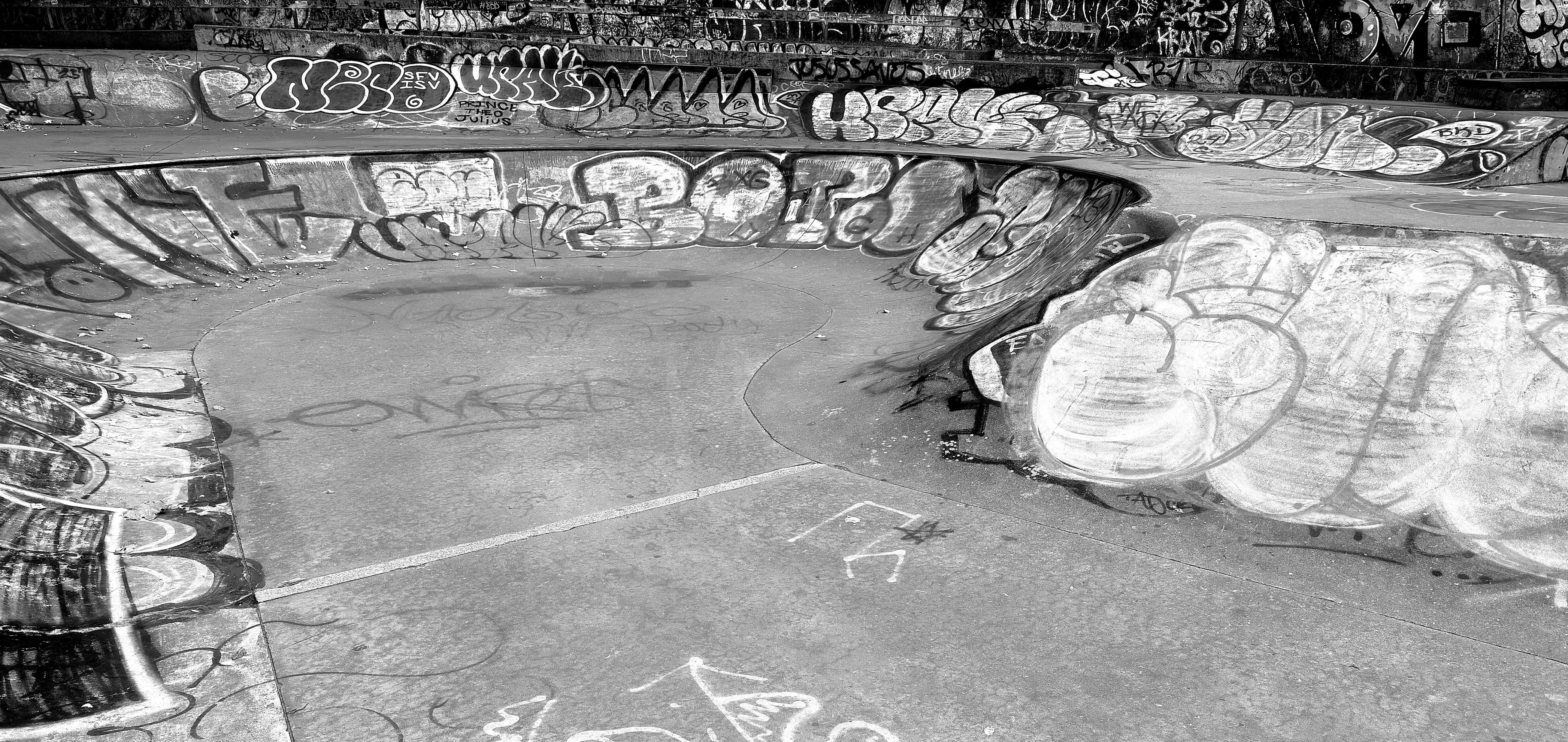 A noir photograph of a gritty urban concrete skate park bowl void of people along River Avenue in the Highbridge neighborhood of the Bronx. The bowl has smooth, curved sides that transition to a flat bottom. There are dozens of graffiti tags ranging from large, bold bubble letters primarily covering the curved walls, to smaller tags and scribbles across the floor and upper perimeter. The upper edges of the park show tiers of seating steps, and they are also heavily covered in darker graffiti.