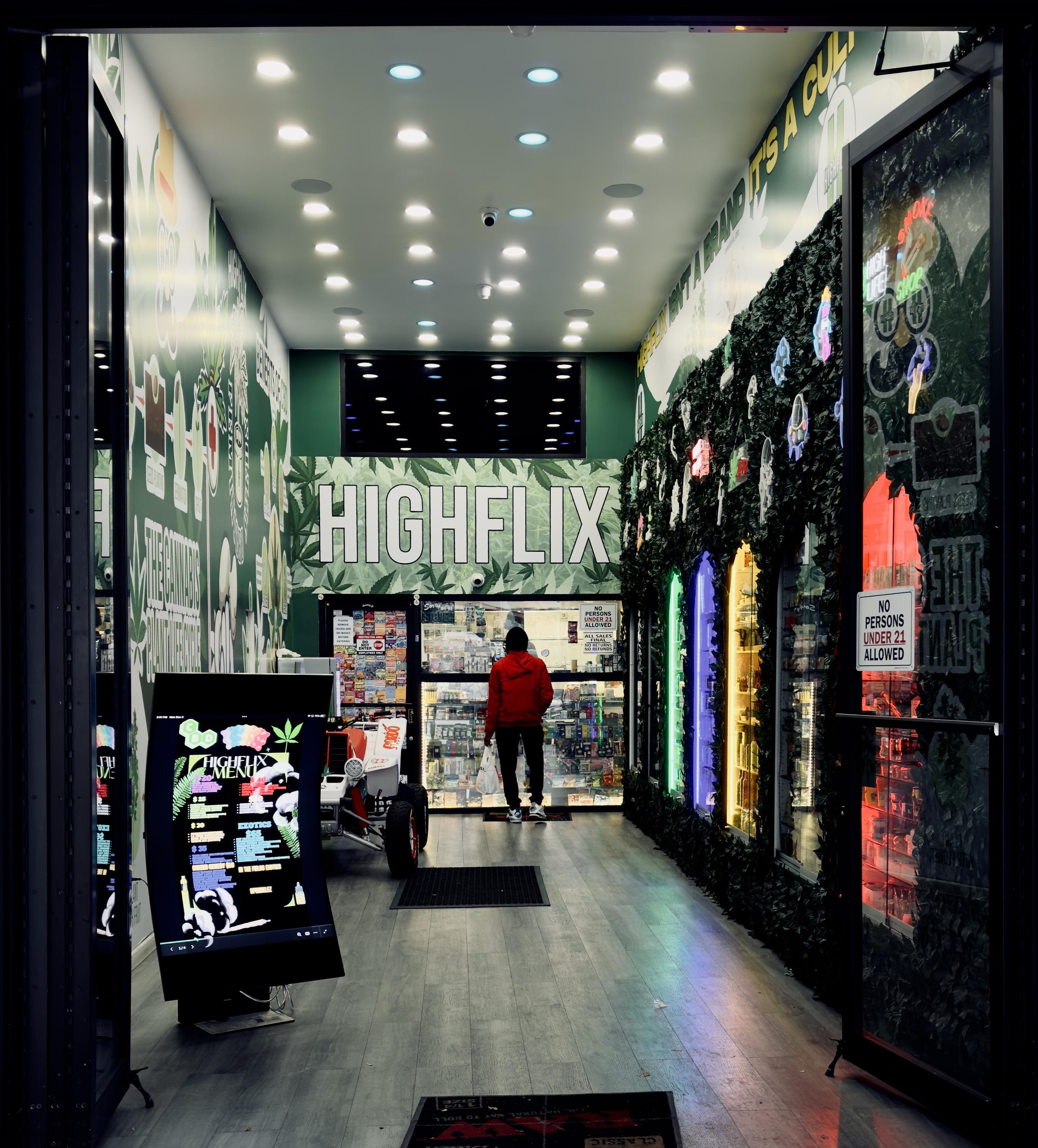A color photograph of the interior entrance to a small cannabis dispensary in the Marble Hill neighborhood of Manhattan. The dispensary is defined by a high-contrast aesthetic, primarily using light/dark green, black, and bright white. The walls feature a graphic pattern with cannabis leaf illustrations against a white background. Dominating the back wall is the dispensary's name, HIGHFLIX, rendered in large, bright white, all-caps lettering. The ceiling is lined with numerous small, circular LED spotlights in pastel colors of cream, blue, pink. Embedded into the right wall are several vertical display windows, each backlit with a different bright neon color: green, blue, yellow, and red. Behind the windows are cannabis products. A sign is visible on the inner surface of the right entrance door, stating, "NO PERSONS UNDER 21 ALLOWED." A customer wearing a red hoodie and black pants stands in the back of the store, facing a set of clear glass windows enclosing the cashier. In the foreground to the left is a tall, vertical digital screen displaying a black-and-white graphic menu titled "HIGHFLIX MENU" with a cannabis leaf logo, resting on a black base. The floor is a light wood laminate, and a dark welcome mat advertising Raw rolling papers lies at the entrance.