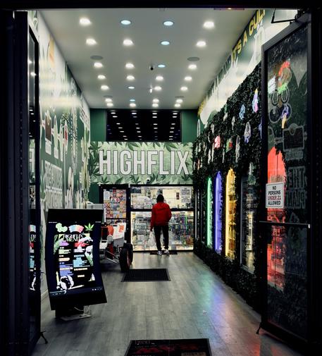 A color photograph of the interior entrance to a small cannabis dispensary in the Marble Hill neighborhood of Manhattan. The dispensary is defined by a high-contrast aesthetic, primarily using light/dark green, black, and bright white. The walls feature a graphic pattern with cannabis leaf illustrations against a white background. Dominating the back wall is the dispensary's name, HIGHFLIX, rendered in large, bright white, all-caps lettering. The ceiling is lined with numerous small, circular LED spotlights in pastel colors of cream, blue, pink. Embedded into the right wall are several vertical display windows, each backlit with a different bright neon color: green, blue, yellow, and red. Behind the windows are cannabis products. A sign is visible on the inner surface of the right entrance door, stating, "NO PERSONS UNDER 21 ALLOWED." A customer wearing a red hoodie and black pants stands in the back of the store, facing a set of clear glass windows enclosing the cashier. In the foreground to the left is a tall, vertical digital screen displaying a black-and-white graphic menu titled "HIGHFLIX MENU" with a cannabis leaf logo, resting on a black base. The floor is a light wood laminate, and a dark welcome mat advertising Raw rolling papers lies at the entrance.