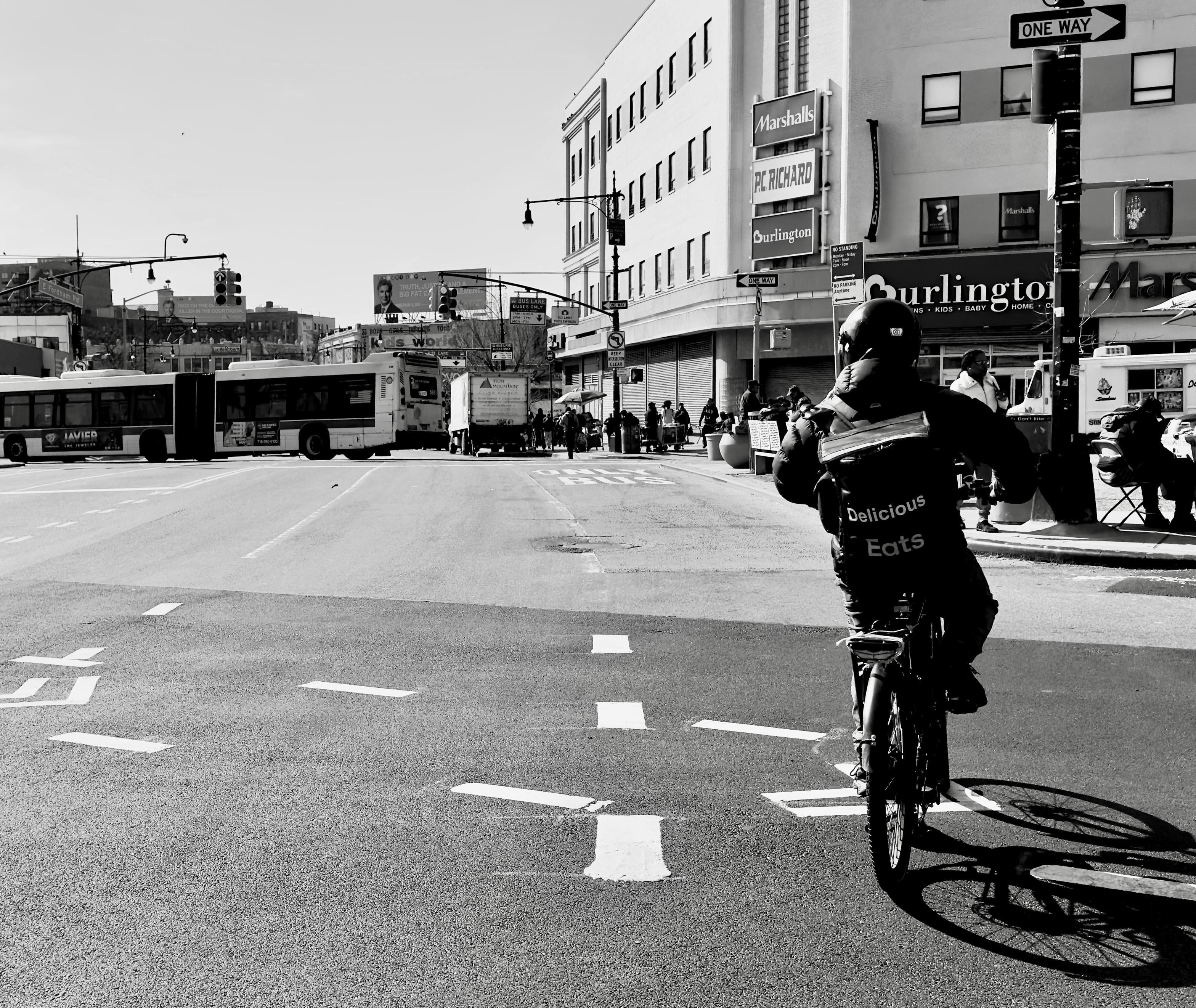 A noir photograph in square aspect ratio of the street scene along the Grand Concourse in the Fordham neighborhood of the Bronx. It is morning on a clear day in early Spring. The focal point of the image captures a cyclist from behind, positioned in the center-right foreground, crossing the very wide city street. The cyclist is wearing a helmet and a jacket with the words "Delicious. Uber Eats." printed prominently on the back. Their bike casts a long, dark shadow diagonally across the street toward the bottom right corner, contrasting sharply with the bright, sunlit asphalt. The street is marked with white dashed lines for lanes and a turning area, and the composition is meant to draw the viewer's eye down the road toward the middle ground. The background features the busy urban landscape of this commercial section of the Bronx. To the left, a large two section bus is turning left out of the frame. Directly ahead and slightly to the right, a two-story building anchors the intersection, displaying large signs for major retail chains, including "Marshalls," "PC RICHARD," and "Burlington." Pedestrians are visible on the sidewalk in front of the stores. A street sign with "ONE WAY" is mounted on a pole to the far right. The strong midday sun creates deep shadows and high contrast, emphasizing the architectural lines of the buildings and the dynamic movement of the street scene. A clear sky defines the horizon.