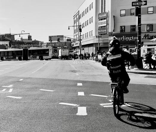 A noir photograph in square aspect ratio of the street scene along the Grand Concourse in the Fordham neighborhood of the Bronx. It is morning on a clear day in early Spring. The focal point of the image captures a cyclist from behind, positioned in the center-right foreground, crossing the very wide city street. The cyclist is wearing a helmet and a jacket with the words "Delicious. Uber Eats." printed prominently on the back. Their bike casts a long, dark shadow diagonally across the street toward the bottom right corner, contrasting sharply with the bright, sunlit asphalt. The street is marked with white dashed lines for lanes and a turning area, and the composition is meant to draw the viewer's eye down the road toward the middle ground. The background features the busy urban landscape of this commercial section of the Bronx. To the left, a large two section bus is turning left out of the frame. Directly ahead and slightly to the right, a two-story building anchors the intersection, displaying large signs for major retail chains, including "Marshalls," "PC RICHARD," and "Burlington." Pedestrians are visible on the sidewalk in front of the stores. A street sign with "ONE WAY" is mounted on a pole to the far right. The strong midday sun creates deep shadows and high contrast, emphasizing the architectural lines of the buildings and the dynamic movement of the street scene. A clear sky defines the horizon.