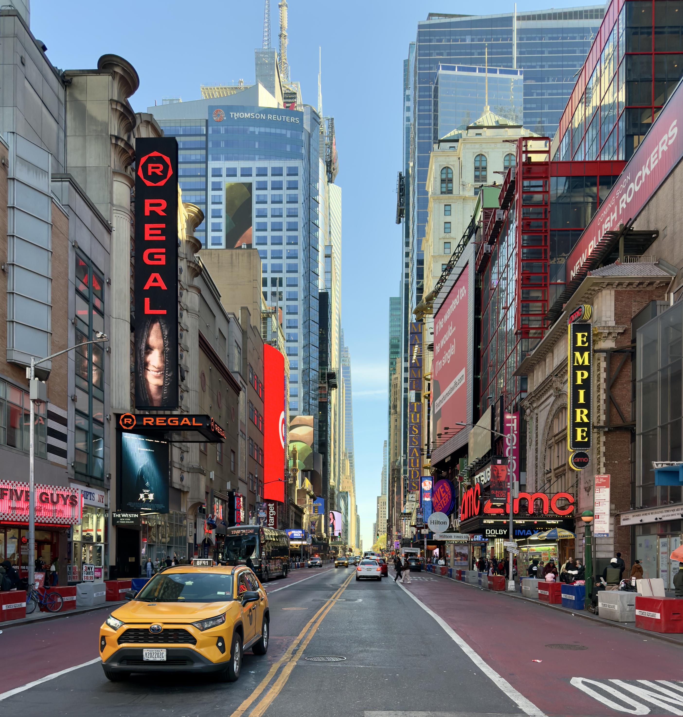 A color photograph of West 42nd Street looking east from 8th Avenue toward Times Square in Manhattan. The street is nearly empty of traffic, with only a taxi van driving toward the camera in the left lane. A few pedestrians are visible on the sidewalks. Large, multi-story buildings with numerous electronic billboards and theater marquees line both sides of the street. On the left, Thomson Reuters tower, and a prominent Regal movie theater sign with vertical lettering and a stylized eye logo is visible. A Five Guys restaurant is also visible on the left, next to the theater. On the right, the Empire Theatre and AMC Empire 25 movie theater buildings are prominent. The overall tone is emphasizing the urban canyon effect of the architecture and signage.