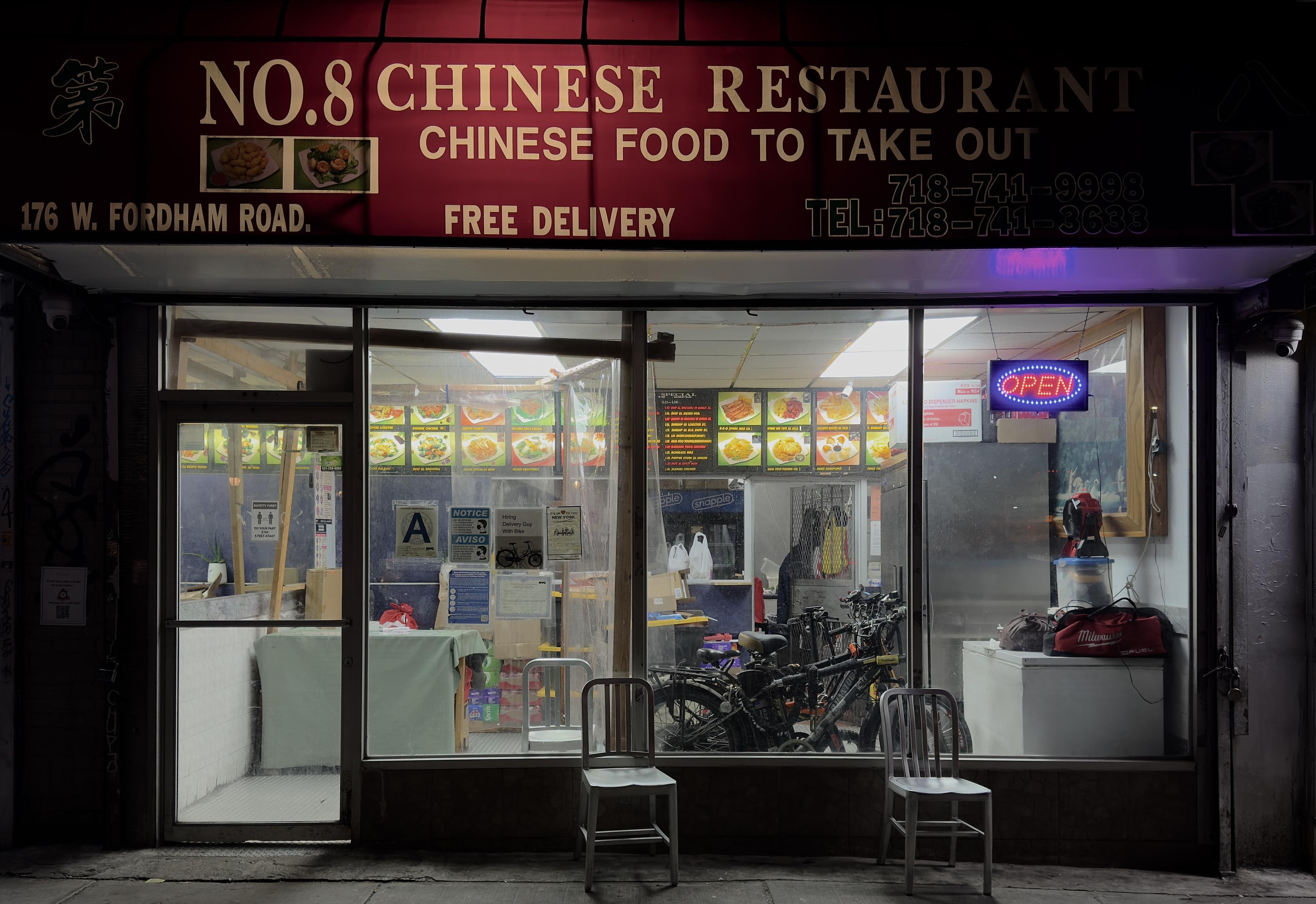 A color photograph in landscape orientation taken outside of a Chinese restaurant late at night. The sign above the storefront is a deep red, with large white and yellowed lettering that reads "NO. 8 CHINESE RESTAURANT" and below it "CHINESE FOOD TO TAKE OUT." Smaller text indicates the address, "176 W. FORDHAM ROAD.", and includes two phone numbers, "TEL: 718-741-9993" and "TEL: 718-741-3633," as well as "FREE DELIVERY." The sign also features a small image of two prepared food dishes. The interior is brightly lit, contrasting sharply with the dark street. Through the large glass windows, several hanging menu boards and displayed photos of food dishes are visible. An "OPEN" neon sign glows in the right window. The interior visible through the glass windows shows the establishment fully. Several delivery bicycles and bags are stored to the right, adjacent to a white refrigerator. Various notices and regulatory signs are posted on the inside of the glass door and windows. Two plain, metal chairs are placed on the sidewalk outside the storefront. The interior lights are casting white light onto the sidewalk of the storefront.