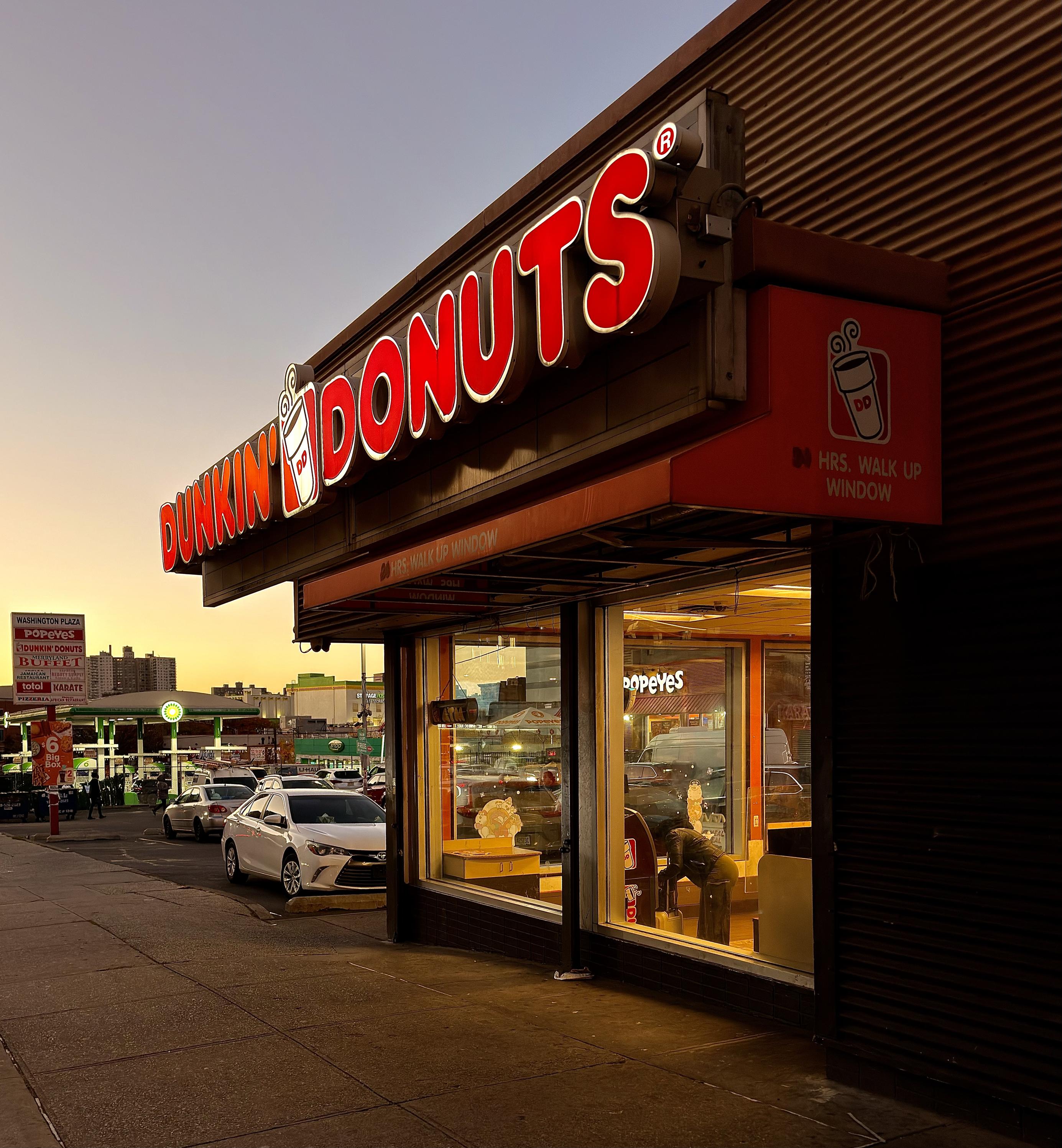 A color, street-level photograph captures the street scene of a brightly lit Dunkin' Donuts storefront at dusk. The large, red-and-white "DUNKIN' DONUTS" sign, featuring a stylized coffee cup logo, dominates the top of the frame and glows against the warm orange and twilight blue sky. Below the main sign, a smaller red sign indicates "HRS. WALK UP WINDOW." Inside the store, warm interior lights illuminate a customer seated at a table near the window. The perspective shows the sidewalk and a few cars parked in the strip mall parking lot, including a white sedan closest to the foreground. In the background to the left, a Popeyes Louisiana Kitchen sign is visible through the windows of the coffee shop and above a gas station canopy.