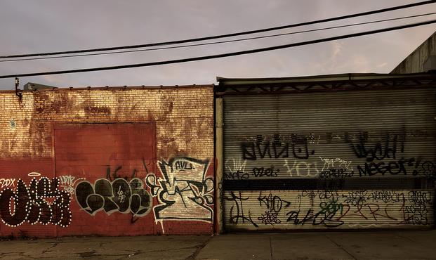 A color, wide-angle photograph in landscape orientation taken at dusk of a weathered defunct warehouse heavily covered in graffiti. The scene is split by two distinct surfaces: on the left is a tan brick wall with a prominent, rectangular rolling gate. The gate and the lower half of the wall has been sloppily painted red. The wall features large, bubble-style graffiti in white and black near the bottom left, alongside other smaller tags. The bricks themselves are stained and aged, with a subtle golden glow from a lamp post light source outside the frame. It is starkly contrasted against the fading natural light of the evening sky. The right half of the image is dominated by a corrugated metal roll-down security gate, which is also extensively covered in small black, white, and red graffiti tags. The metal surface reflects the dim, gray-purple light of the cloudy dusk sky, which is visible across the top of the image. Three sets of utility wires run horizontally across the upper third of the frame, visually separating the sky from the building structure below. The ground in the foreground is a dark, rough pavement sidewalk. The overall mood is dark, gritty, melancholic, and urban.