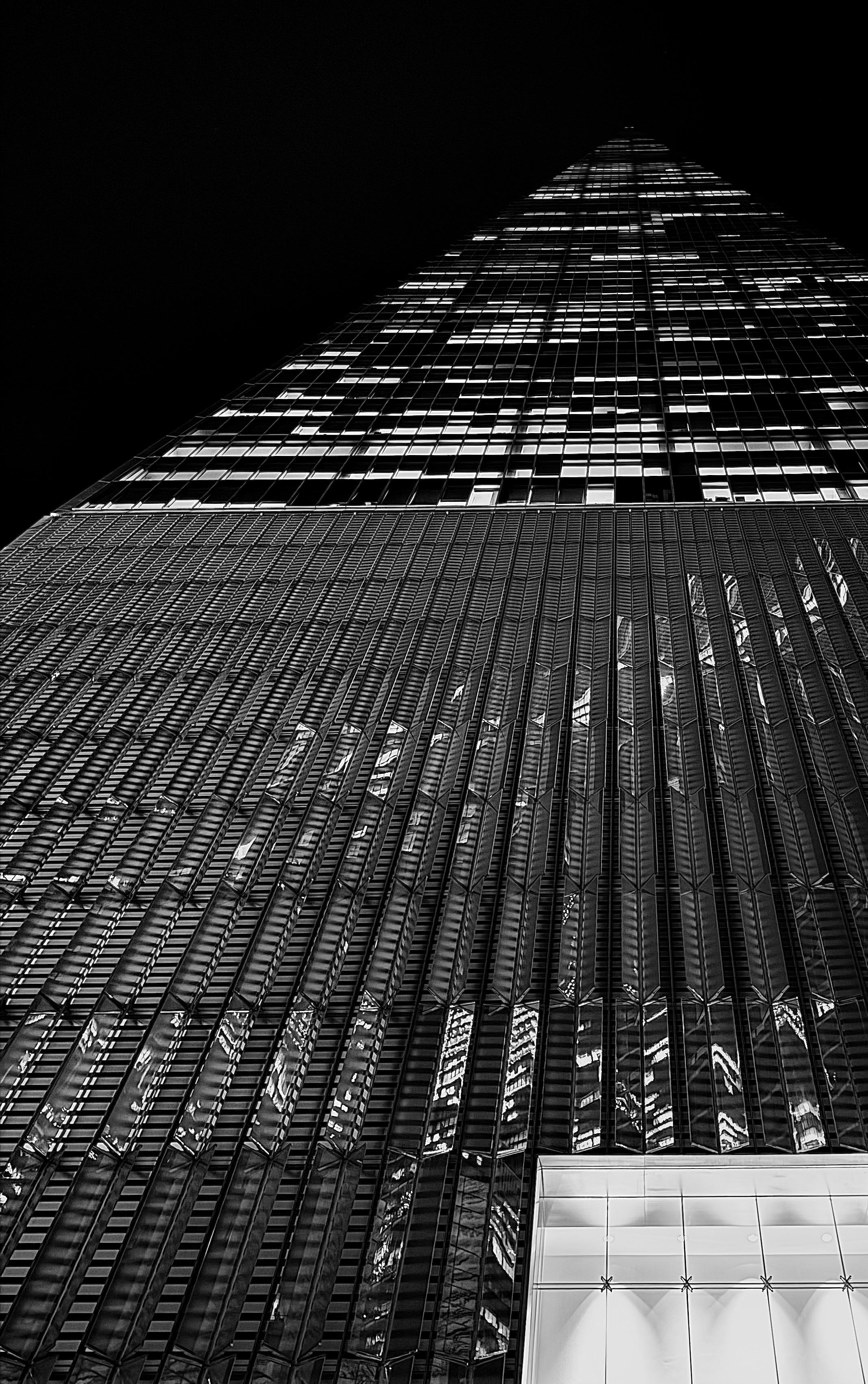 A low-angle, noir photograph of the One World Trade Center tower in New York City taken at night. The composition looks sharply upward, emphasizing the immense height and tapering geometry of the skyscraper. The building dominates the frame against a completely black sky. The lower two-thirds of the image show the tower's base, illuminated entrance, and the initial tiers of its reflective glass, characterized by vertical fins and sharp architectural lines. These sections reflect distorted, bright lights from the interior and surrounding buildings, creating a textured, almost abstract pattern of light and shadow that runs along the glass surface. The upper third of the image is dedicated to the tower's ascent, where the structure narrows toward its pinpoint shaped apex. In this section, the illuminated office windows create a striking grid pattern, with many windows displaying bright squares of light that stand out against the otherwise dark glass and deep black background.