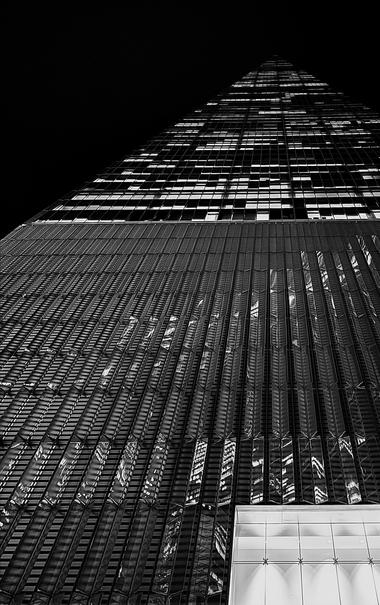 A low-angle, noir photograph of the One World Trade Center tower in New York City taken at night. The composition looks sharply upward, emphasizing the immense height and tapering geometry of the skyscraper. The building dominates the frame against a completely black sky. The lower two-thirds of the image show the tower's base, illuminated entrance, and the initial tiers of its reflective glass, characterized by vertical fins and sharp architectural lines. These sections reflect distorted, bright lights from the interior and surrounding buildings, creating a textured, almost abstract pattern of light and shadow that runs along the glass surface. The upper third of the image is dedicated to the tower's ascent, where the structure narrows toward its pinpoint shaped apex. In this section, the illuminated office windows create a striking grid pattern, with many windows displaying bright squares of light that stand out against the otherwise dark glass and deep black background.