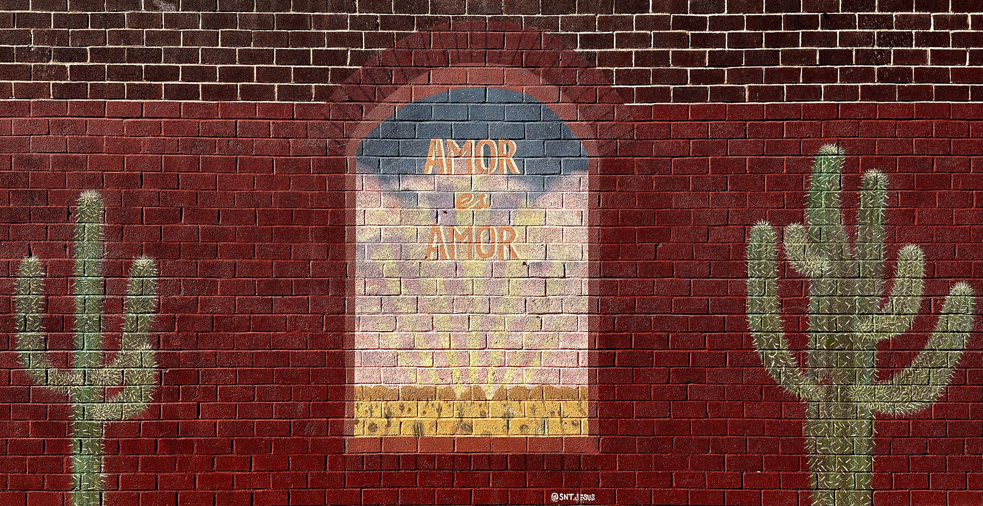 A color, wide-format landscape photograph of a mural painted on a brick wall taken on a sunny morning. The wall itself is a deep, uniform red-brick color with lighter white mortar lines, giving it a classic masonry look. The lower 3/4ths of the wall has been painted maroon red, and the left side is slightly faded, The central focus is a vertically oriented, arched window painted onto the wall, within it appears a soft, sunny scene. The arch frames a gradient of light blue at the top, fading into yellow and light pink in the middle, and ending with a band of dark yellow/orange at the bottom, and depicting a desert landscape filled with cactus and shrubs viewed through a window. Across this glowing area, the phrase "AMOR ES AMOR" (Spanish for "Love is Love") is painted in large, slightly stylized orange and gold block capital letters, stacked one above the other. On noth the left and right sides are two large, stylized paintings of cacti. The cacti are depicted using a light green paint that allows the red brick texture to show through, making them look almost like illuminated silhouettes. Both cacti have a central trunk and multiple curving arms. A small, white signature tag reads ‘@SNTJESUS’ is visible centered at the bottom edge of the arched window painting.