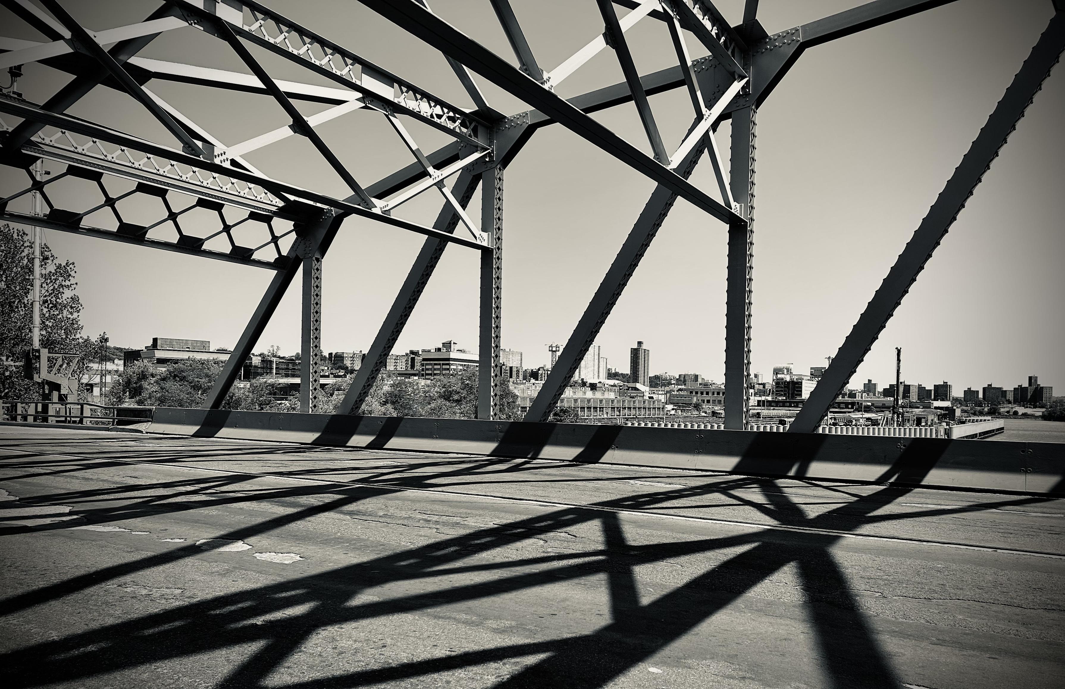 A monochrome photograph of the University Heights Bridge between Manhattan and the Bronx, taken from the pedestrian walking path. The scene is dominated by the bridge's massive, angular steel truss structure, which creates strong geometric patterns. The dark steel members, cast in bright sunlight, form dramatic, intersecting diagonals and triangles that frame the upper left and center of the shot. The harsh sunlight creates long, sharply defined black shadows of the structure on the cracked, asphalt road. In the background, a hazy skyline of low-rise buildings and industrial structures is visible across the Harlem River.