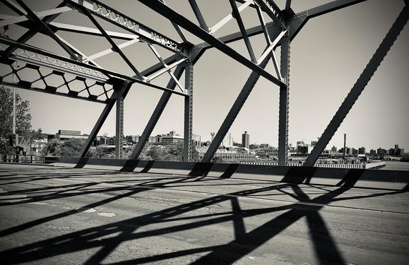 A monochrome photograph of the University Heights Bridge between Manhattan and the Bronx, taken from the pedestrian walking path. The scene is dominated by the bridge's massive, angular steel truss structure, which creates strong geometric patterns. The dark steel members, cast in bright sunlight, form dramatic, intersecting diagonals and triangles that frame the upper left and center of the shot. The harsh sunlight creates long, sharply defined black shadows of the structure on the cracked, asphalt road. In the background, a hazy skyline of low-rise buildings and industrial structures is visible across the Harlem River.
