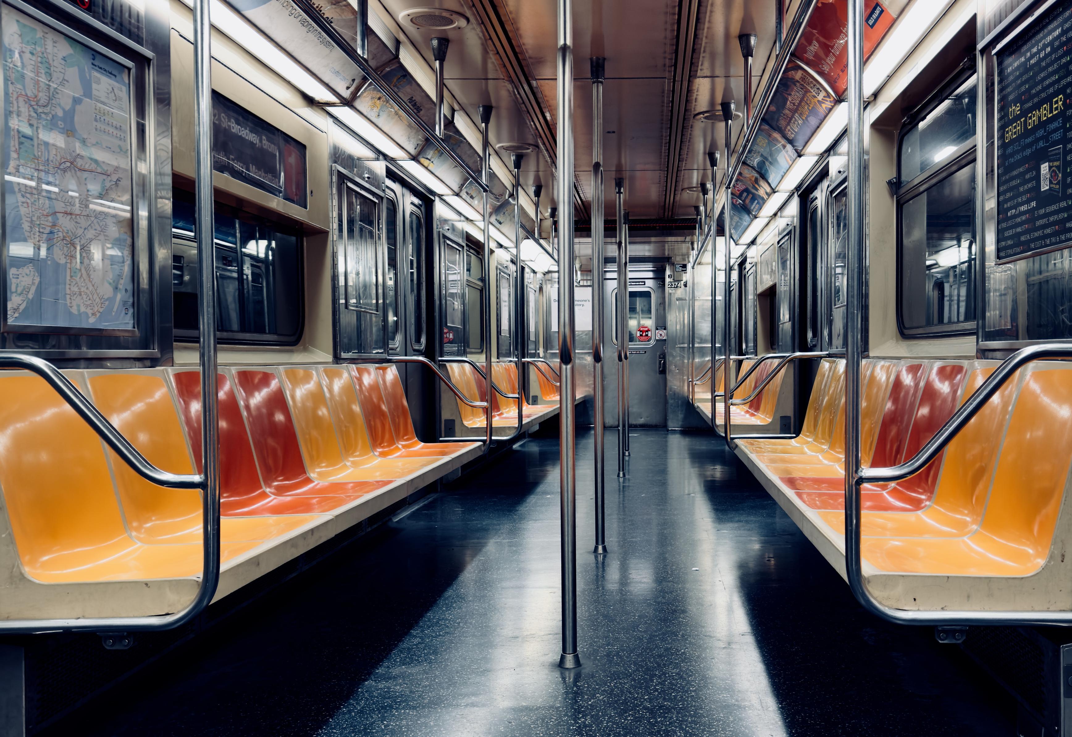 A color, low-angle, wide photograph of an empty New York City subway car. The scene captures the car from the center aisle, looking straight towards the opposite end, creating a sense of perspective. The seating runs along both side walls, featuring a gradient of colors from a deep reddish-orange near the backrests to a brighter, almost golden yellow on the seat fronts, which are illuminated by overhead strip lights. Several vertical, polished stainless steel grab bars run down the center aisle. The floor is a dark, reflective gray. On the left side, a large framed subway map is visible behind glass, while on the right, a smaller advertisement for "The Great Gambler is visible. The car is running through the tunnel so no light is shining in through the windows.