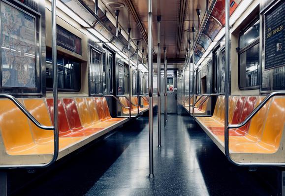A color, low-angle, wide photograph of an empty New York City subway car. The scene captures the car from the center aisle, looking straight towards the opposite end, creating a sense of perspective. The seating runs along both side walls, featuring a gradient of colors from a deep reddish-orange near the backrests to a brighter, almost golden yellow on the seat fronts, which are illuminated by overhead strip lights. Several vertical, polished stainless steel grab bars run down the center aisle. The floor is a dark, reflective gray. On the left side, a large framed subway map is visible behind glass, while on the right, a smaller advertisement for "The Great Gambler is visible. The car is running through the tunnel so no light is shining in through the windows.