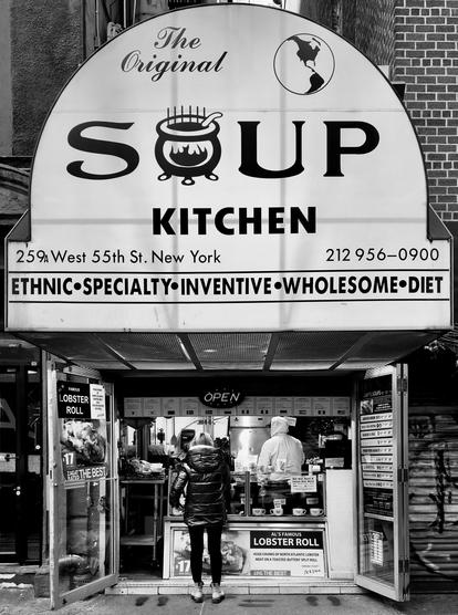 A noir photograph of the street scene at the storefront of The Original Soup Kitchen located in Manhattan. The main sign is a large, arched white panel with bold black text. The top section reads "The Original" with a small logo featuring a globe and map outline, while the central text prominently displays the word "SOUP," with the letter 'O' replaced by an illustration of a steaming cauldron. Below this is the word "KITCHEN," and underneath, the address and phone number: "259A West 55th St. New York 212 956-0900." A horizontal white banner directly beneath the main sign lists descriptive food categories in centered, capitalized text: "ETHNIC • SPECIALTY • INVENTIVE • WHOLESOME • DIET." The storefront below is open, revealing the small open sidewalk service counter area inside where an employee wearing a white hooded winter coat are working. A lone customer, seen from the back and wearing a dark puffer jacket, dark pants, and sneakers stands just inside the doorway looking at the menu boards above the counter. The space is narrow, with display materials and signs visible, including one on the left advertising a "FAMOUS LOBSTER ROLL" with a picture of the item. The building facade is framed by the brick structure. On the right side of the frame a rolling shutter gate is partially visible, closed and covered in graffiti tags.