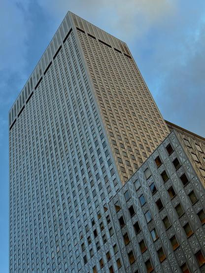 A color photograph in portrait orientation that captures the dramatic upward view of a large, modern skyscraper in midtown Manhattan against a partly cloudy blue sky. The building occupies most of the frame, towering diagonally from the lower right toward the upper left. The main structure is characterized by its sheer verticality and a highly textured, grid-like facade composed of numerous small, repeated rectangular windows and metal panels. The material is a silvery-gray muted metallic color, reflecting the ambient light unevenly.
