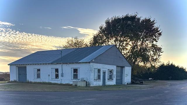 A color photograph in landscape orientation of an old, single-story, white masonry building. It is situated next to a paved road in a rural Kansas setting. The building has a prominent, steep-pitched metal roof that is a light blue-gray. The white painted concrete walls are weathered, featuring five windows and two large garage bay doors, one on the far left and one on the right. The sky above is a bright blue, with wispy, streaky cirrocumulus clouds catching the warm, golden light of the low evening sun. The background is dominated by a large, dark-leafed deciduous tree immediately behind the building, whose foliage is silhouetted against the bright sky, its leaves casting a dark shadow across the scene. To the far right, there are smaller, dense evergreen shrubs.