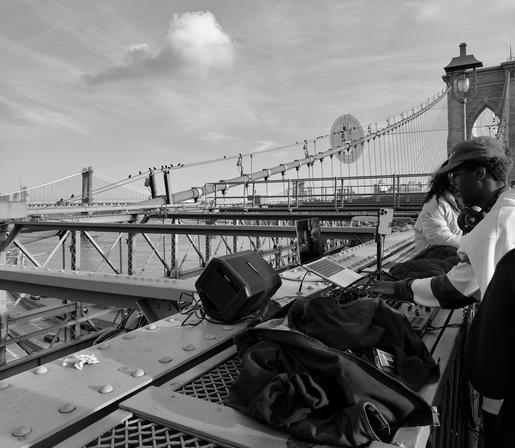 A noir photograph taken along the Brooklyn Bridge in New York City on a partly cloudy summer day. The primary focus is on two individuals near a makeshift live streaming setup consisting of a large speaker, a laptop, an iPhone 16 Pro Max, and sound mixing gear. One person, wearing a baseball cap and headphones hanging around their neck, is actively engaging with the equipment on the right side of the frame. The bridge has distinct latticework and a metal deck. Beyond the immediate foreground activity, the photo shows the expansive scale of the structure. The suspension cables and towers of the Brooklyn Bridge dominate the background, stretching into the distance and framing the scene. Further in the background the Manhattan Bridge is visible spanning the East River.