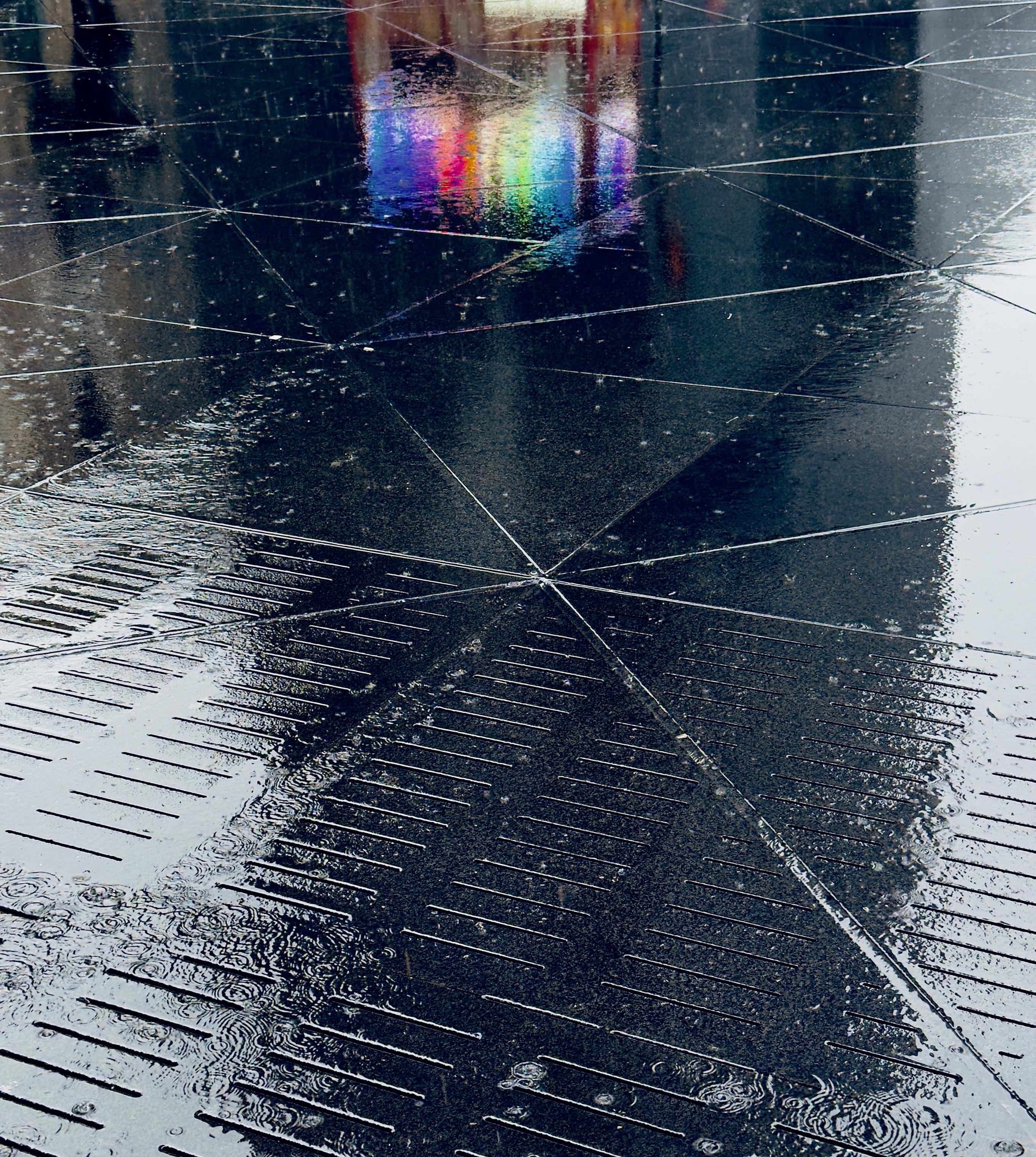 A color photograph in portrait orientation captures a moody, dramatic view of a rain covered sidewalk outside Moynihan Train Hall in New York City. The sidewalk has been transformed into a mirror by the downpour. The foreground is dominated by the wet, dark paving stones, where the rain creates a scattering of small ripples and glistening reflections. The texture of the sidewalk features a pattern of horizontal grooves in the lower half that transition into larger, smooth, dark tiles crisscrossed by intersecting, bright grout lines. The heavy rain enhances the contrast. The upper portion of the frame has a splash of color in an otherwise monochromatic scene from the lights of a street food vendor cart that is not directly visible. This reflection appears as a distinct, rectangular prism of light and color, dominated by a vibrant rainbow effect.