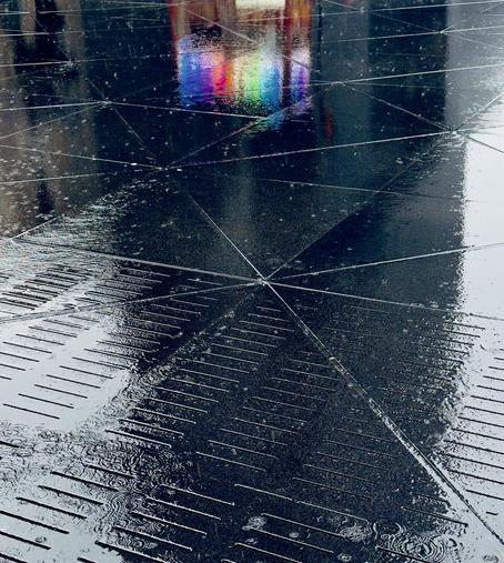 A color photograph in portrait orientation captures a moody, dramatic view of a rain covered sidewalk outside Moynihan Train Hall in New York City. The sidewalk has been transformed into a mirror by the downpour. The foreground is dominated by the wet, dark paving stones, where the rain creates a scattering of small ripples and glistening reflections. The texture of the sidewalk features a pattern of horizontal grooves in the lower half that transition into larger, smooth, dark tiles crisscrossed by intersecting, bright grout lines. The heavy rain enhances the contrast. The upper portion of the frame has a splash of color in an otherwise monochromatic scene from the lights of a street food vendor cart that is not directly visible. This reflection appears as a distinct, rectangular prism of light and color, dominated by a vibrant rainbow effect.
