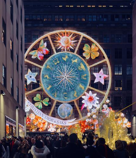 A color, portrait oriented photograph taken at night of the Saks Fifth Avenue holiday store display from the winter of 2023 in midtown Manhattan. It features a colossal, illuminated Ferris wheel-like structure designed in collaboration with Christian Dior. The main structure dominates the center frame and is a massive golden wheel with a turquoise-blue central disc. This disc is decorated with intricate golden symbols representing the twelve signs of the zodiac, radiating out from a central hub. The golden frame of the wheel is adorned with brilliantly illuminated, oversized decorative elements, including a vibrant orange, pink, and red butterfly, large four-leaf clovers, bright pink and purple stars, and white and orange flowers. The entire structure is held aloft by a scaffolding that also supports a banner across the bottom reading "CHRISTIAN DIOR & SAKS." Below this main wheel, the lower part of the display features a dense, intricate arrangement of sparkling gold lights shaped into swirling forms, resembling golden foliage or abstract figures. The display is set between two tall, dark building facades, with numerous windows glowing yellow in the background. The street level is crowded with a large number of people looking up and taking photos of the installation.
