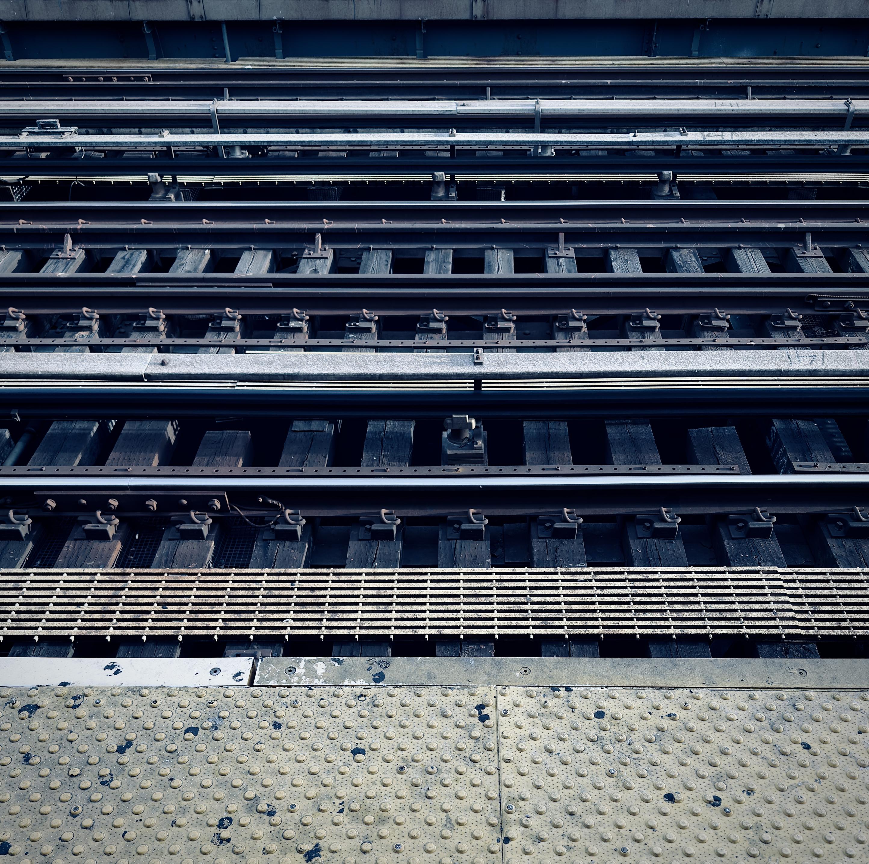 A color photograph capturing a view of multiple sets of elevated subway tracks receding into the distance at the Jackson Avenue elevated subway station in the Woodstock neighborhood of the Bronx. It is an overcast morning so the scene is desaturated from the cool, muted lighting. In the foreground, the concrete edge of the station platform is visible, featuring a prominent band of heavily textured yellow/beige warning surface with circular, raised bumps. Immediately beyond the platform edge is a wide, metal grid drainage grate, showing signs of wear and rust. Three distinct trackways are visible, each composed of dark, worn wooden ties laid perpendicularly across dark steel beams.