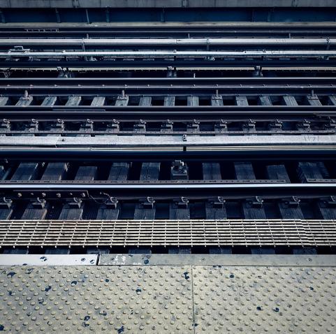A color photograph capturing a view of multiple sets of elevated subway tracks receding into the distance at the Jackson Avenue elevated subway station in the Woodstock neighborhood of the Bronx. It is an overcast morning so the scene is desaturated from the cool, muted lighting. In the foreground, the concrete edge of the station platform is visible, featuring a prominent band of heavily textured yellow/beige warning surface with circular, raised bumps. Immediately beyond the platform edge is a wide, metal grid drainage grate, showing signs of wear and rust. Three distinct trackways are visible, each composed of dark, worn wooden ties laid perpendicularly across dark steel beams.