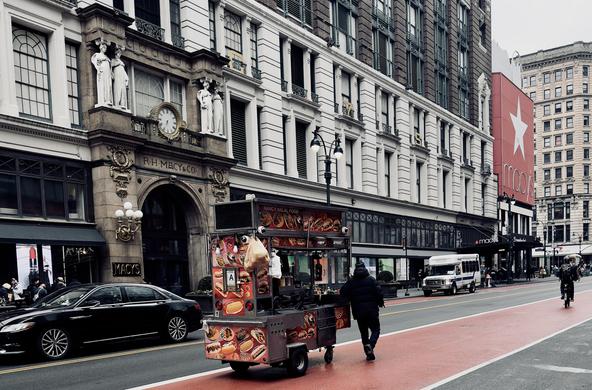 A color photograph of the street scene on 34th Street in front of the historic Macy's Herald Square department store. The foreground is dominated by a food cart being walked down the street by a person in dark clothing facing away from the viewer. To the left, a black sedan is driving towards the left side of the frame. The background is defined by the imposing architecture of Macy's: a large, multi-story building featuring white stone, prominent vertical windows, and elaborate gilded accents around the main entrance. Above the entryway, a large circular clock is visible, flanked by statues. The building's name is subtly integrated into the façade's design, reading "R.H. Macy & Co." Beyond the Macy's building, the street continues into the mid-ground where rows of taller commercial buildings line the right side of the street. Further down the street at the intersection, the iconic red and white Macy's star logo is visible on the side of the building facade. The street features multiple lanes, including the a red-painted lane in the foreground. In the far right of the frame a cyclist is riding away from the camera.