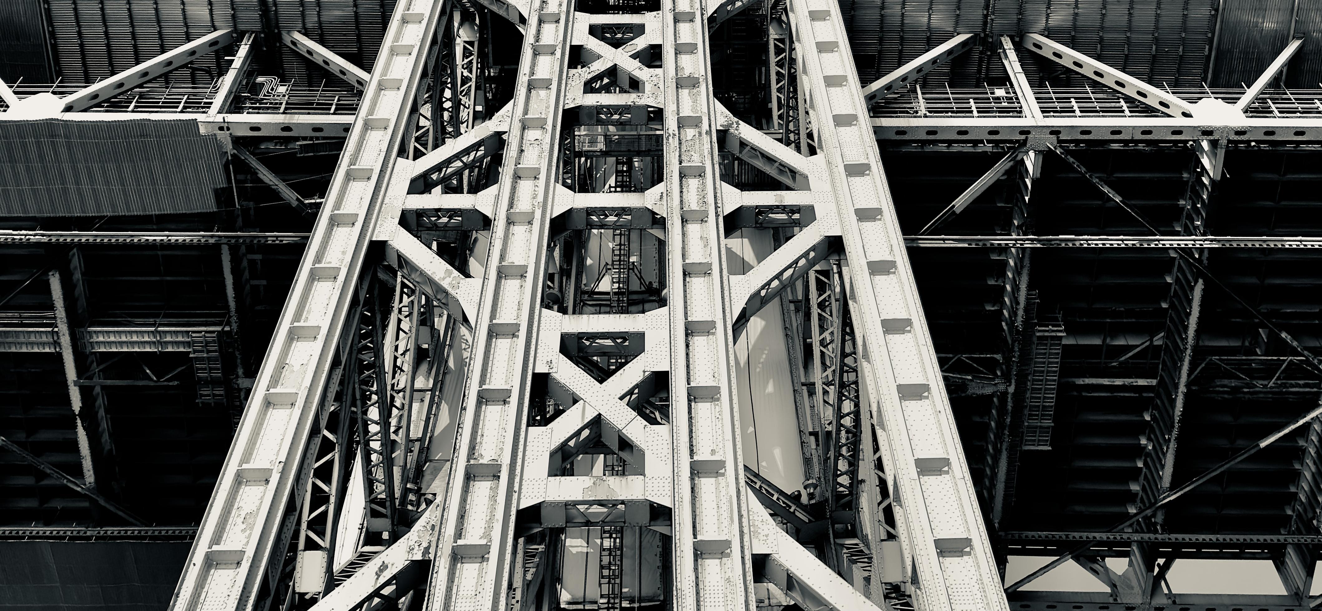 A noir photograph in letterbox format of the monumental structural framework of the George Washington Bridge on a sunny Autumn morning. The view is from directly underneath, looking straight up into the towering lattice of the massive steel column. Visible within the structure are maintenance ladders. The bridge fills the entire frame.